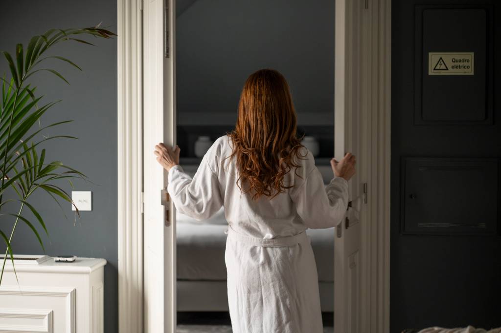 2024_Nuvem-179 Mulher de roupãp abre uma porta de madeira branca num dos quartos do hotel