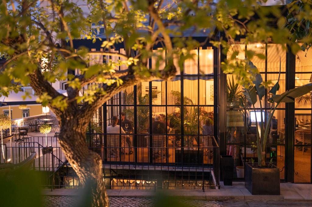 2024_Nuvem-181 A imagem mostra o restaurante do hotel emoldurado por uma árvore do jardim, com fachada de vidro que deixa ver o interior com luzes acesas e mesas ocupadas ao anoitecer