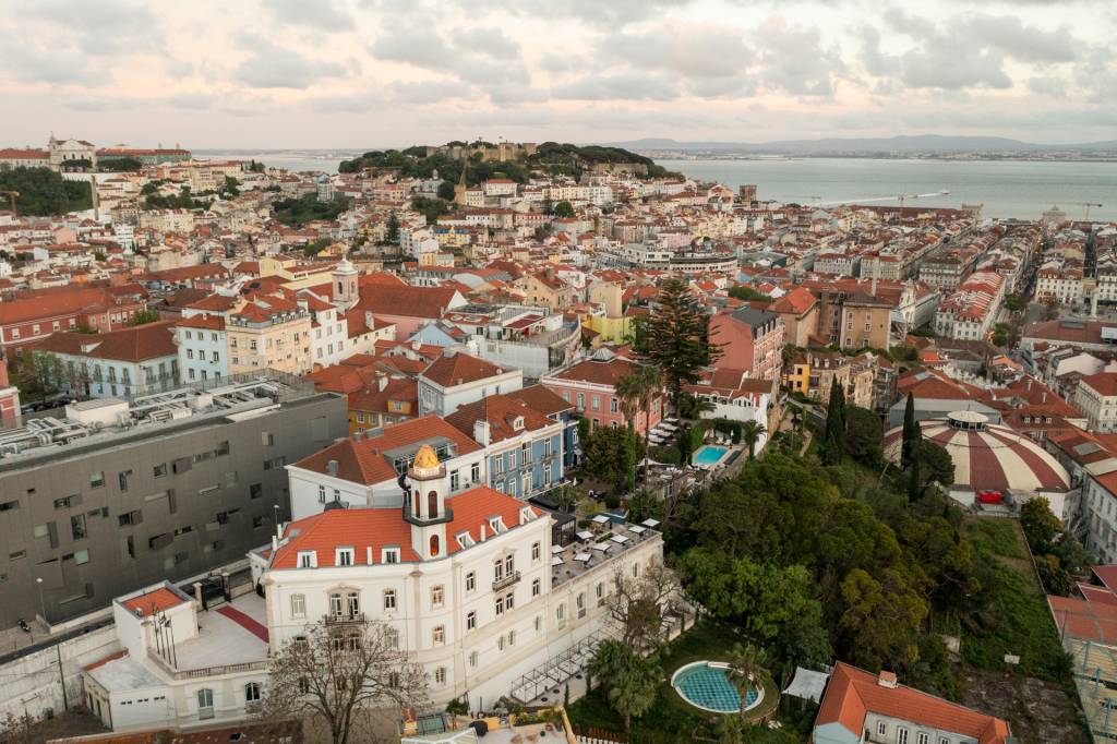 2024_Nuvem-187 Imagem aérea mostra o casario colorido de Lisboa com destaque para um edifício branco com uma torre e uma piscina redonda, onde fica o hotel Torel Palace Lisboa