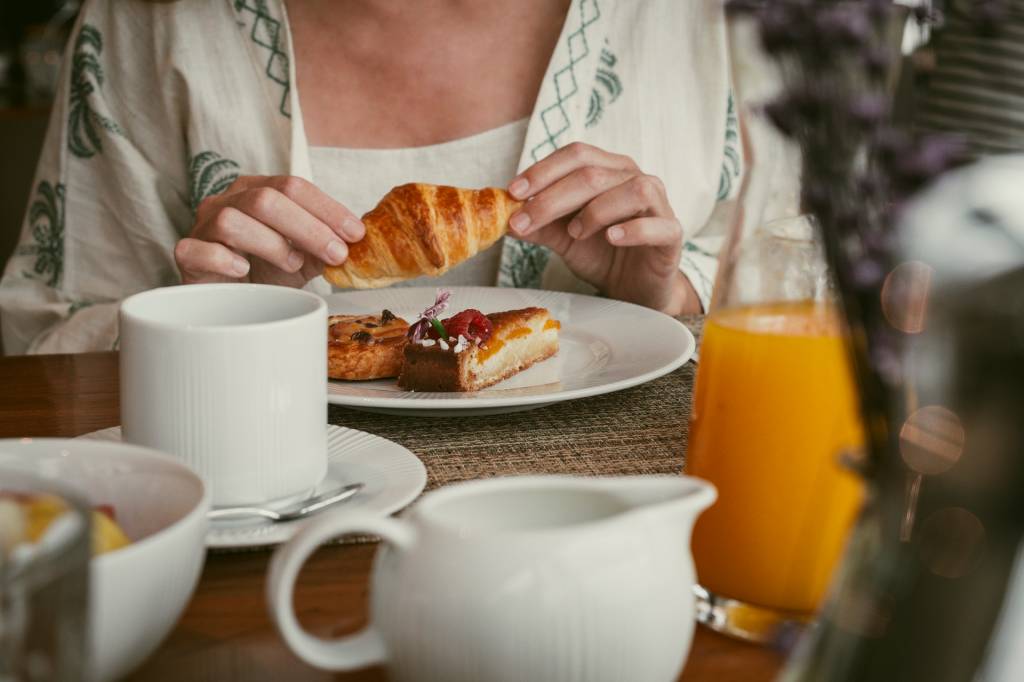 Detalhe de uma mesa de café da manhã do hotel: hóspede a segurar um croissant, com um copo de suco de laranja à frente