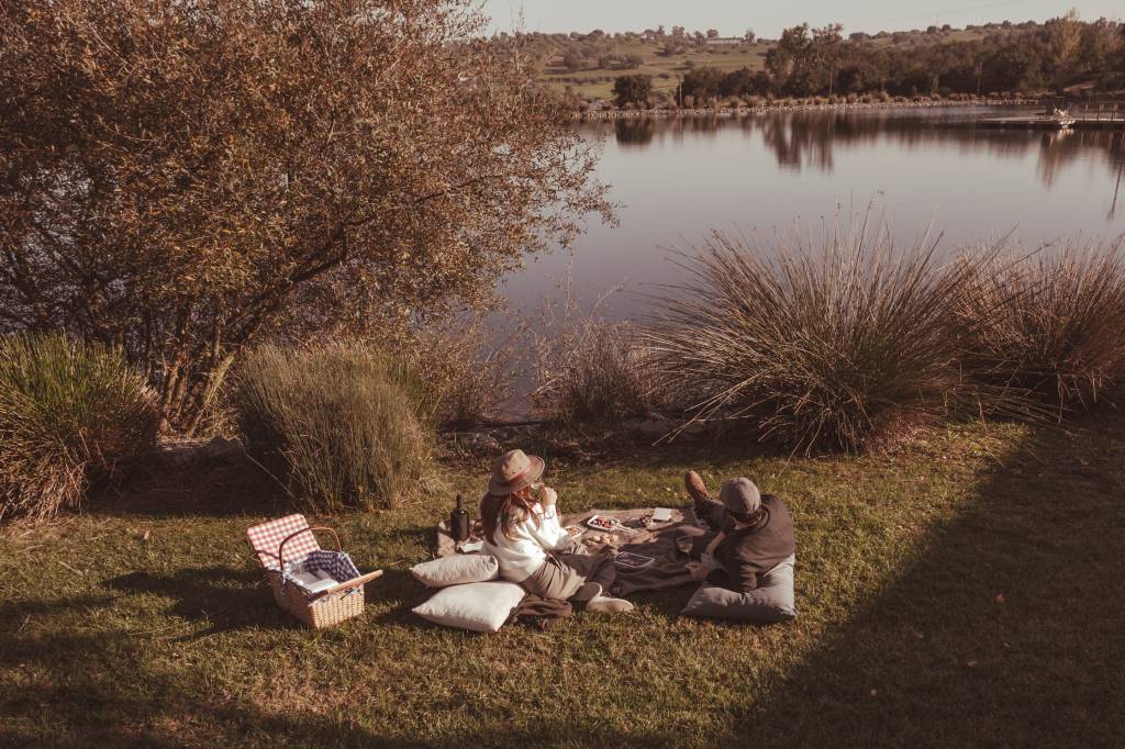 A imagem mostra um casal fazendo piquenique na beira do lago, com uma cesta recheada de guloseimas da região e uma garrafa de vinho