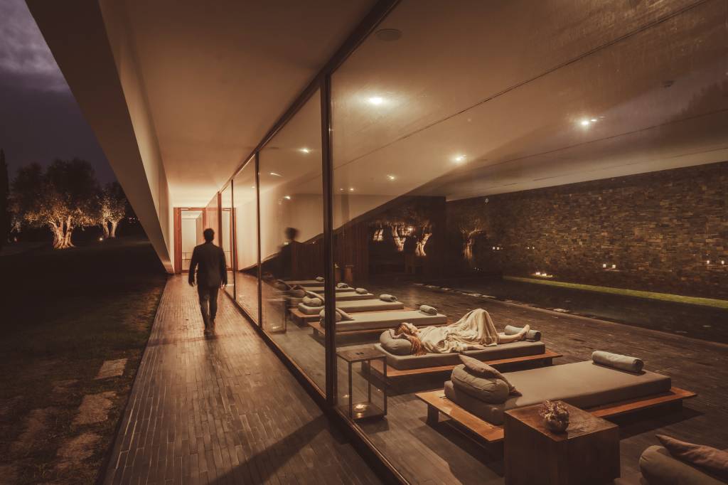 A imagem mostra o spa do hotel, com uma pessoa a caminhar do lado de fora e, do lado de dentro, espreguiçadeiras com os hóspedes em volta da piscina aquecida