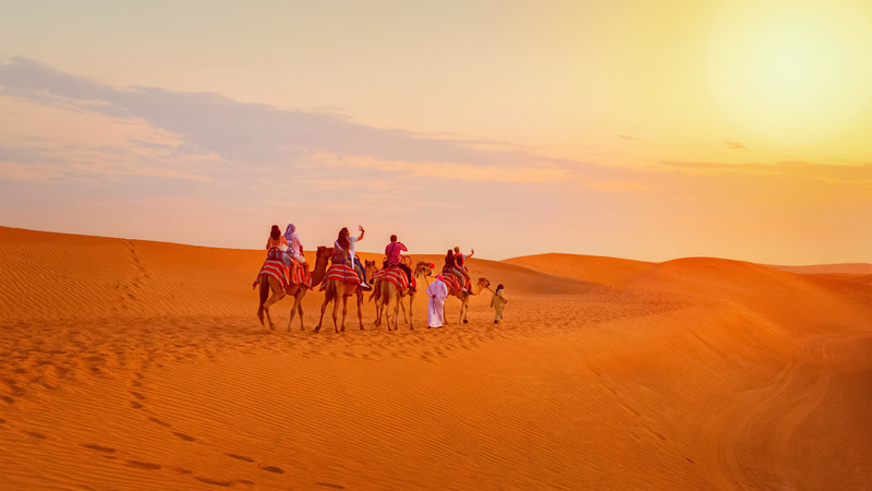 Caravan with group of tourists riding camels through Dubai desert during safari adventure – People exploring sand dunes at sunset in UAE O deserto se transforma em cenário para um passeio único, onde tradição e luxo se encontram