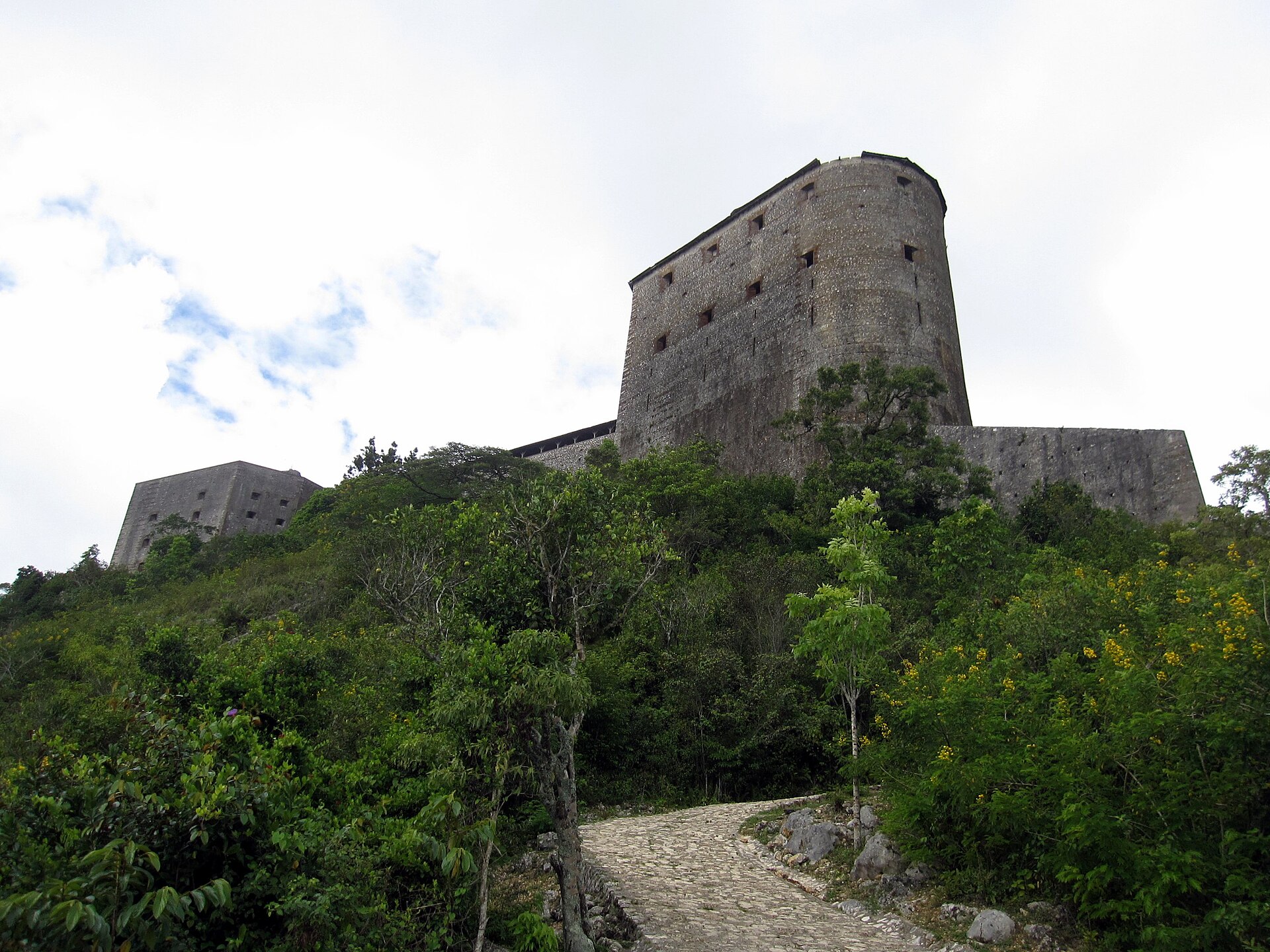 Fortaleza de pedra cinza imponente no topo de uma colina verdejante, com uma estrada de paralelep&iacute;pedos serpenteando em primeiro plano, sob um c&eacute;u nublado