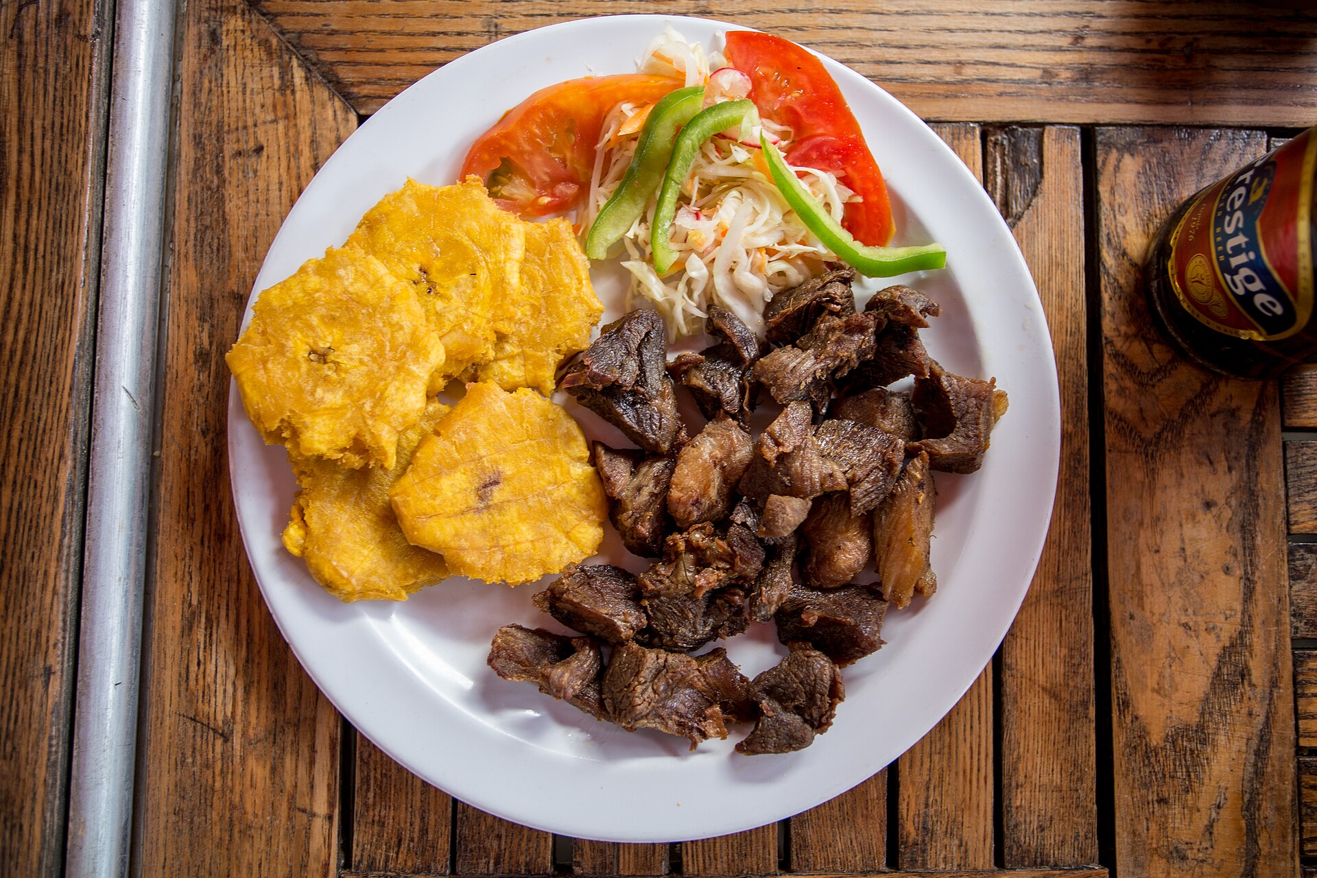 Prato com carne frita em cubos, tostones de banana-da-terra e salada de repolho com tomate e piment&atilde;o verde, sobre mesa de madeira r&uacute;stica, com garrafa de cerveja Prestige ao lado