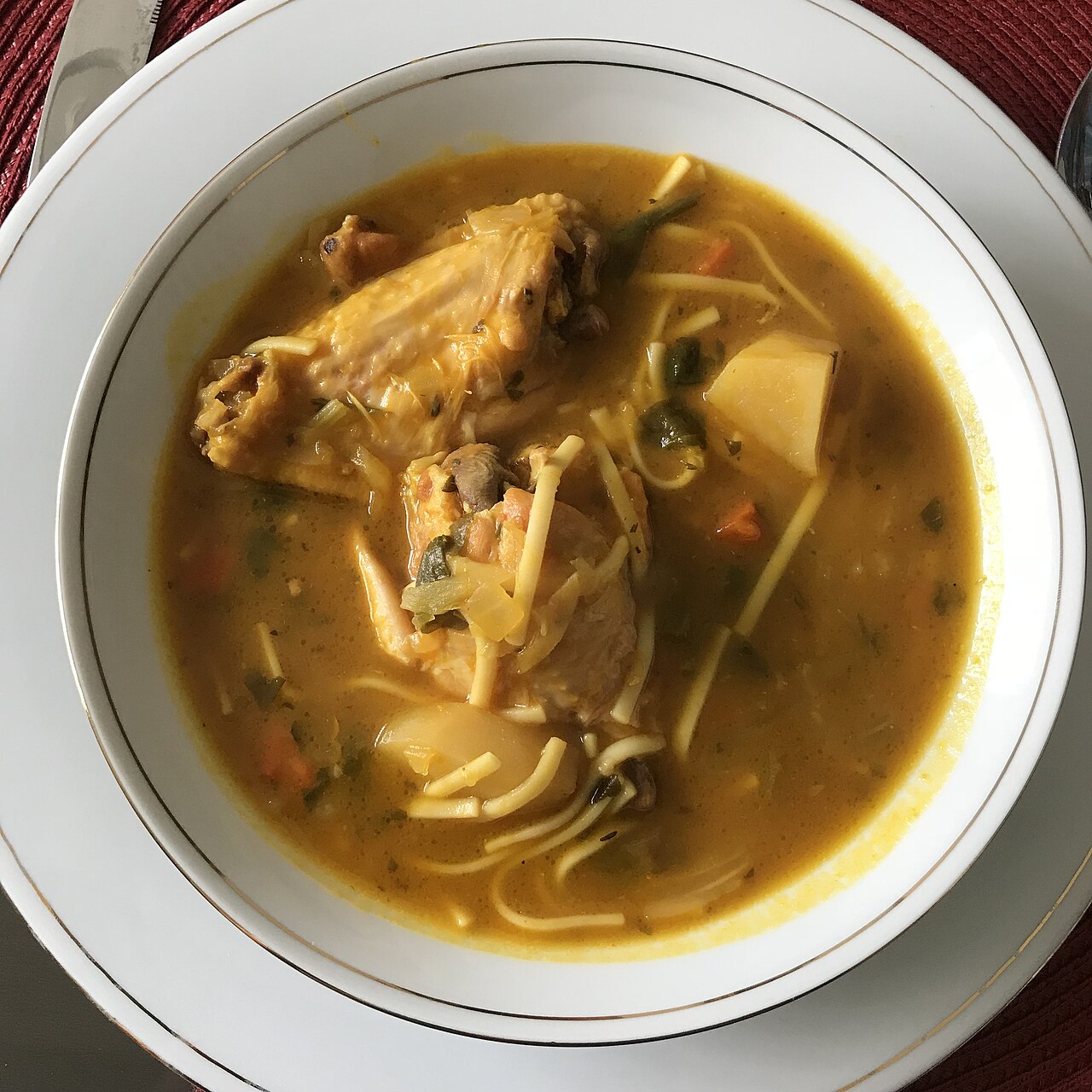 Sopa de galinha com macarr&atilde;o, batata, cenoura e folhas verdes em caldo amarelo, servida em tigela branca com borda dourada sobre prato branco