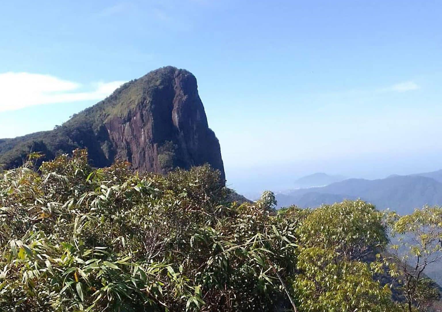 Montanha rochosa e &iacute;ngreme, coberta por vegeta&ccedil;&atilde;o verde no topo, sob c&eacute;u azul claro. Em primeiro plano, arbustos e folhagens densas. Ao fundo, outras montanhas e um vale com n&eacute;voa.