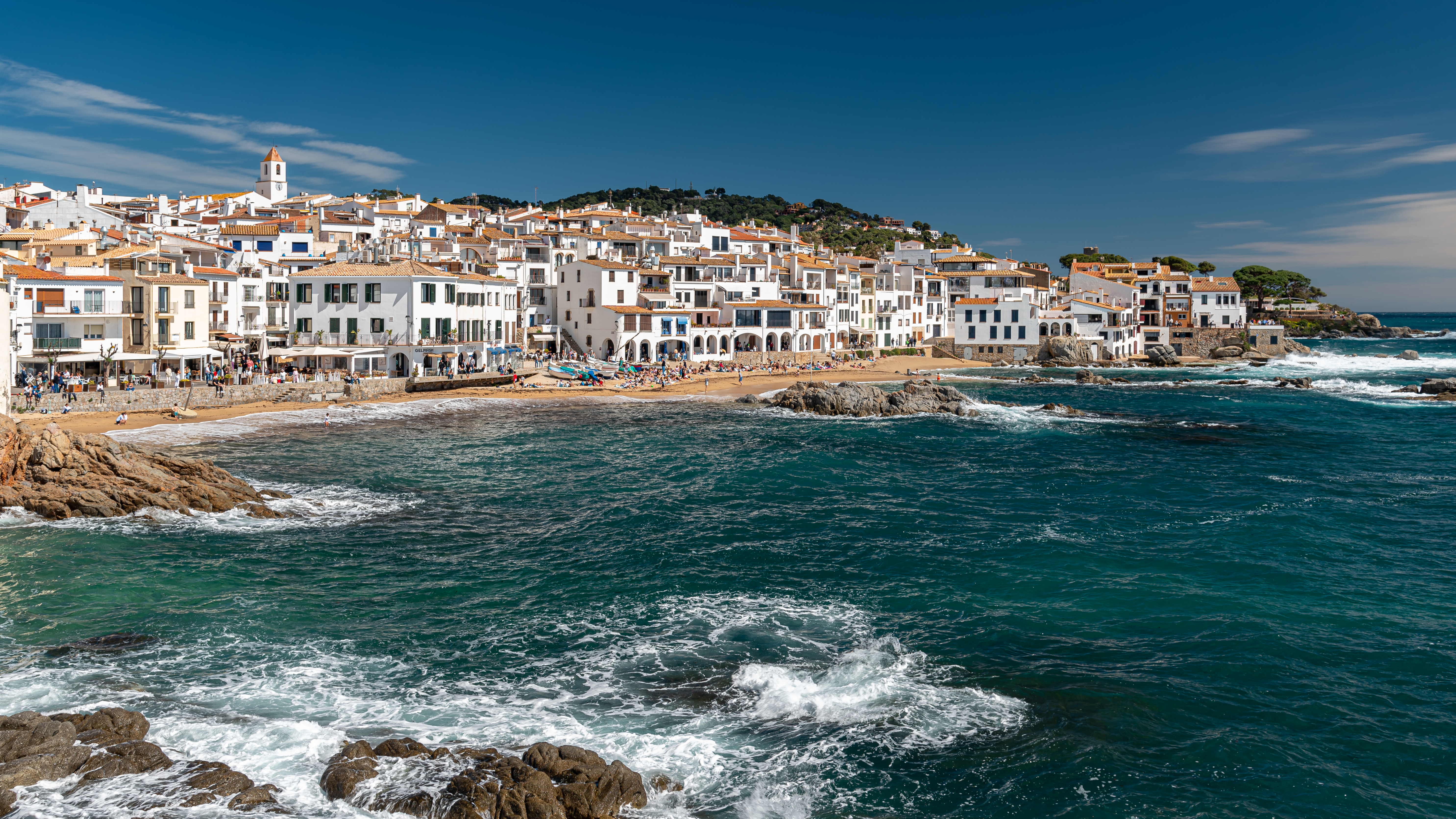 Vila costeira com casas brancas e telhados de terracota, subindo uma colina verde. Uma praia de areia dourada com pessoas e barcos coloridos se estende ao longo da costa, banhada por um mar azul-turquesa com ondas quebrando em rochas escuras. O céu é azul claro, com poucas nuvens. Uma torre de igreja branca se destaca no topo da colina. Foto sob licença CC BY 2.0
