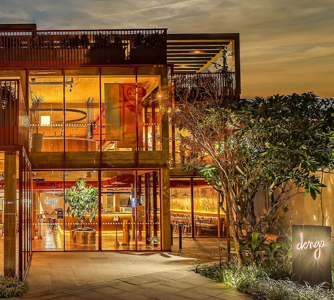 Fachada de um restaurante moderno com grandes janelas de vidro, iluminação quente e um letreiro neon "dengo" à direita, sob um céu alaranjado de fim de tarde