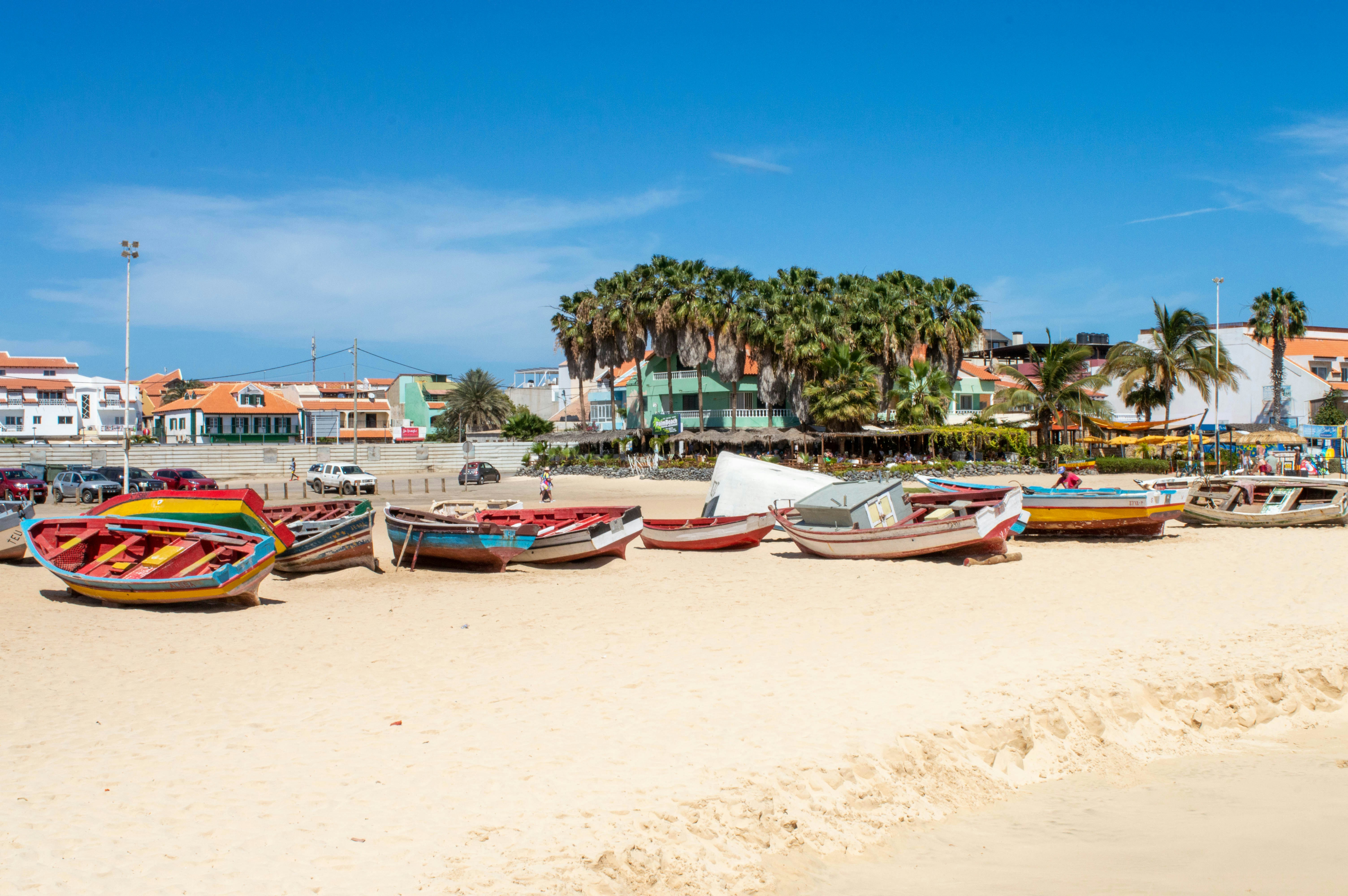 Barcos de pesca coloridos na areia clara de uma praia, com palmeiras e edifícios coloridos ao fundo sob céu azul