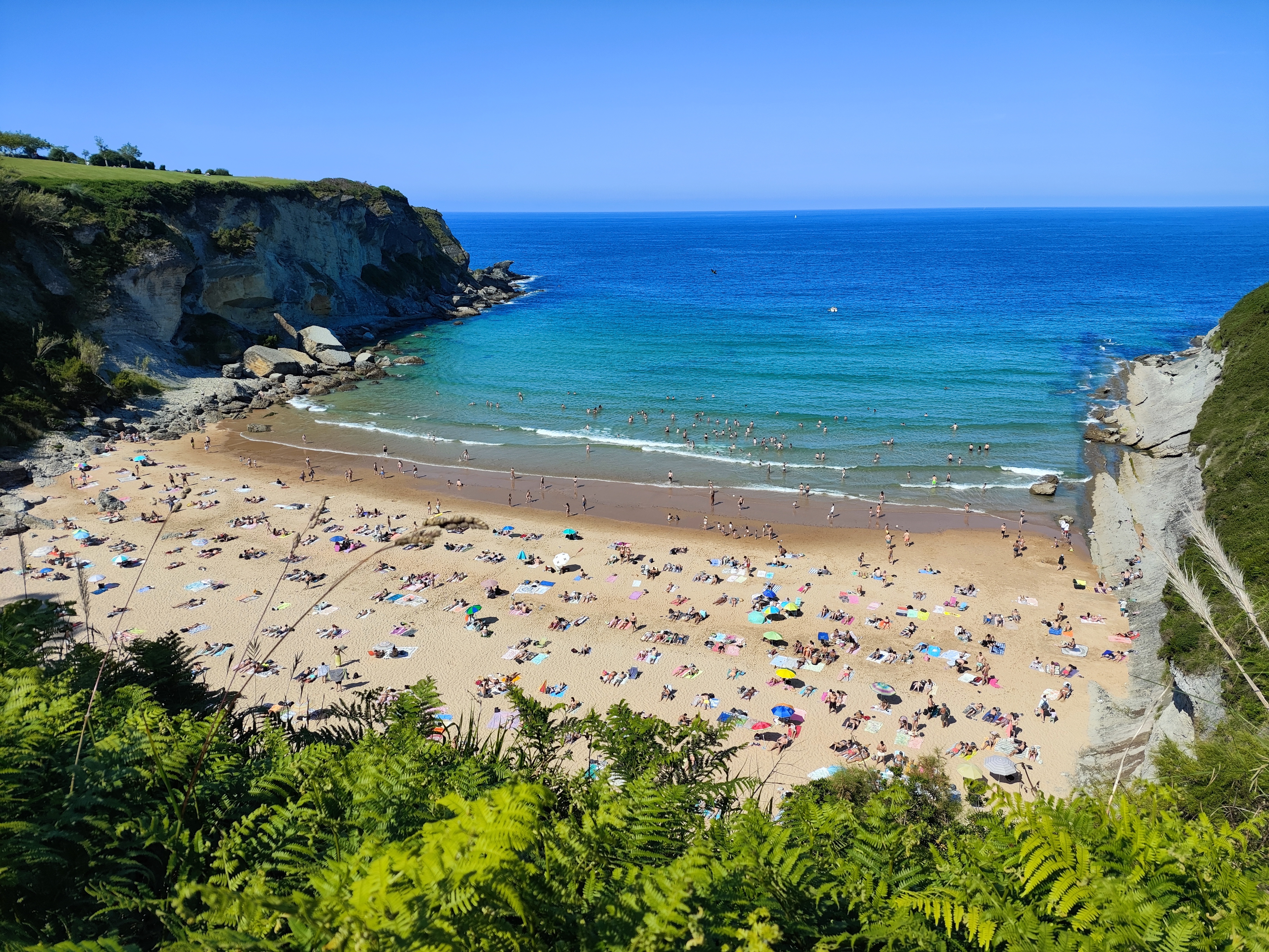 Vista aérea de uma praia lotada, com areia clara e mar azul-turquesa. Pessoas tomam sol em toalhas coloridas e nadam na água. A praia é cercada por falésias rochosas e vegetação verde exuberante, sob um céu azul sem nuvens. Foto sob licença CC BY-SA 4.0