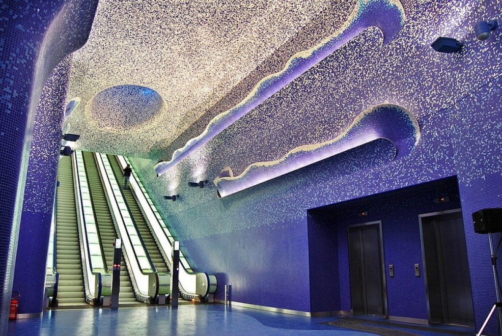 Interior de uma estação de metrô moderna com paredes e teto em mosaico azul e branco, simulando um céu estrelado. Três escadas rolantes sobem à esquerda, com uma pessoa subindo na do meio. À direita, duas portas de elevador escuras se destacam na parede azul. O chão é azul, e o teto tem formas onduladas iluminadas em roxo, criando um efeito futurista