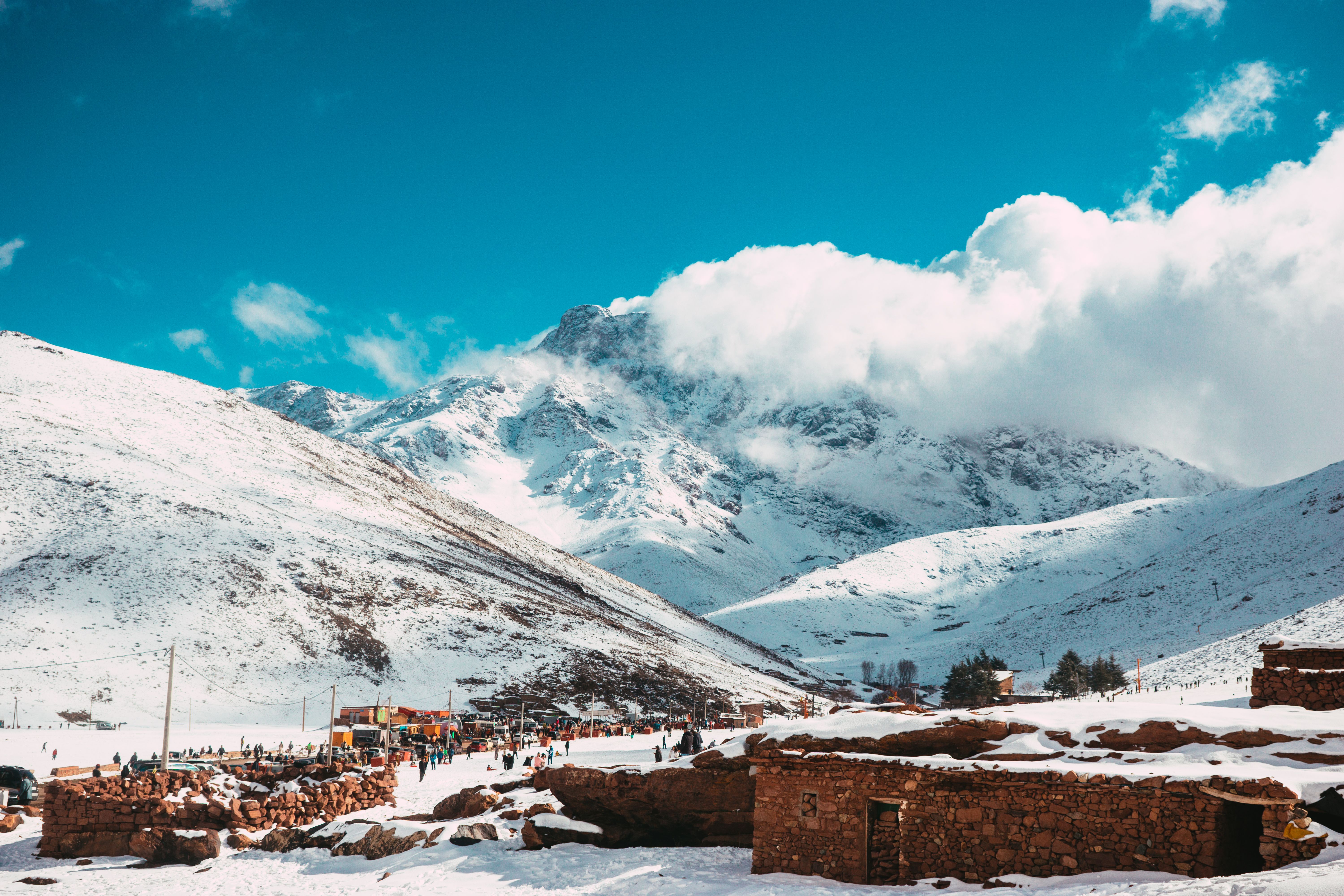 Montanhas nevadas sob um c&eacute;u azul vibrante com nuvens brancas. No vale, um vilarejo com casas de pedra e muitas pessoas caminhando na neve, algumas perto de barracas coloridas. O cen&aacute;rio &eacute; de inverno, com picos cobertos de neve e um clima frio.