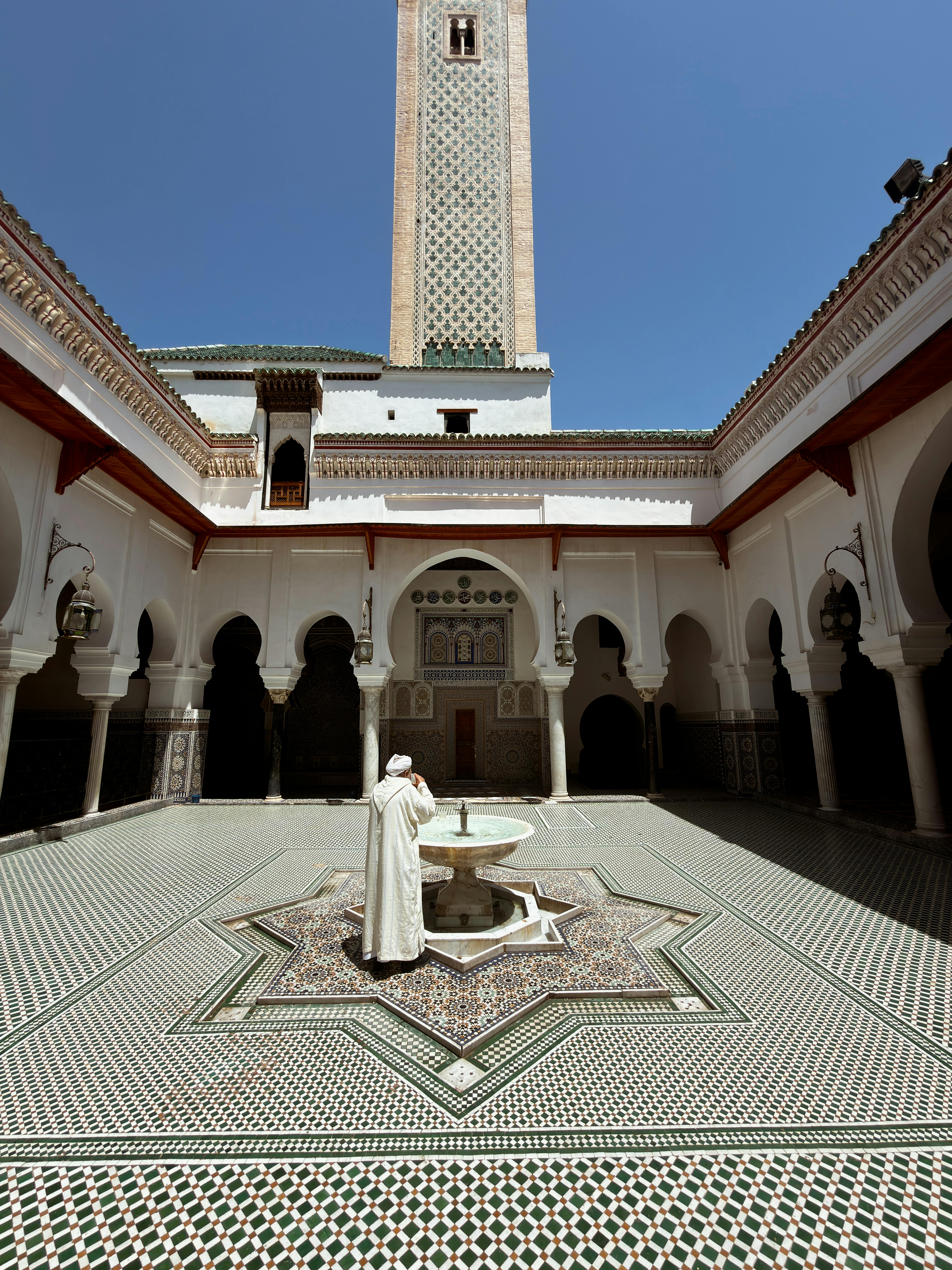 Homem de t&uacute;nica branca e turbante, de costas, em p&aacute;tio de mesquita com fonte central. O ch&atilde;o tem mosaicos verdes e brancos, e ao fundo ergue-se um minarete decorado sob c&eacute;u azul
