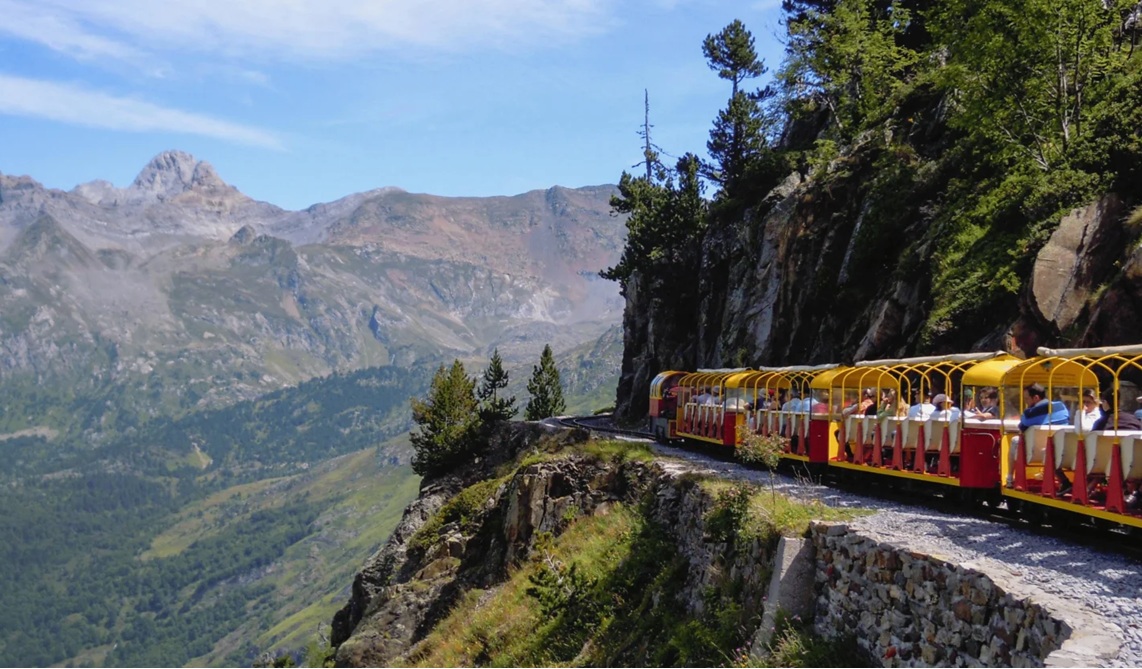Um trem turístico vermelho e amarelo com passageiros percorre uma trilha estreita na encosta de uma montanha rochosa, com um vale verdejante e picos cinzentos ao fundo sob um céu azul