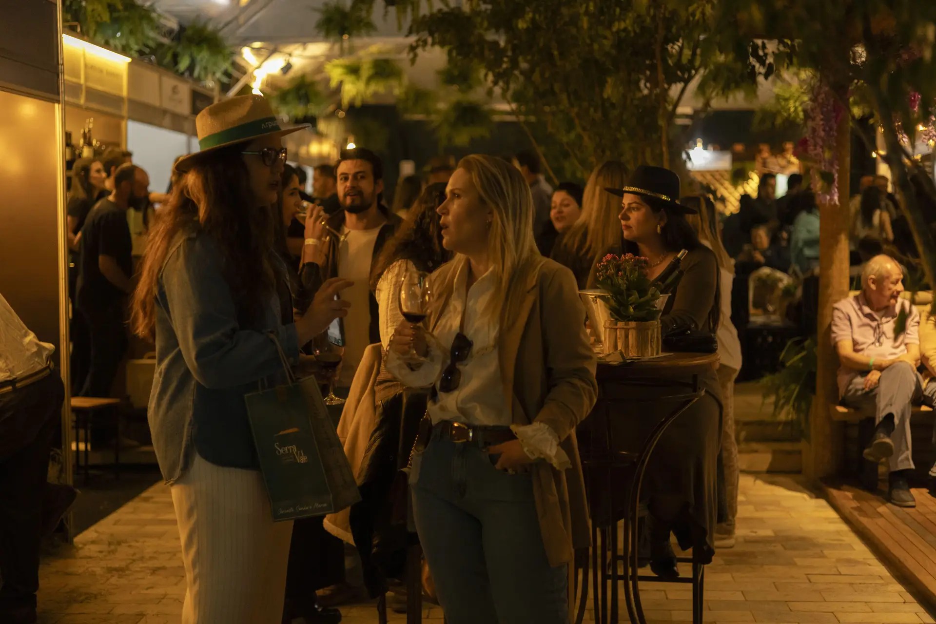 Duas mulheres conversam em um evento noturno. Uma usa chapéu de palha, camisa jeans e calça clara, segurando uma sacola verde. A outra, de blazer bege e calça jeans, segura uma taça de vinho tinto. Ao fundo, pessoas interagem em ambiente iluminado por luzes quentes e vegetação pendente