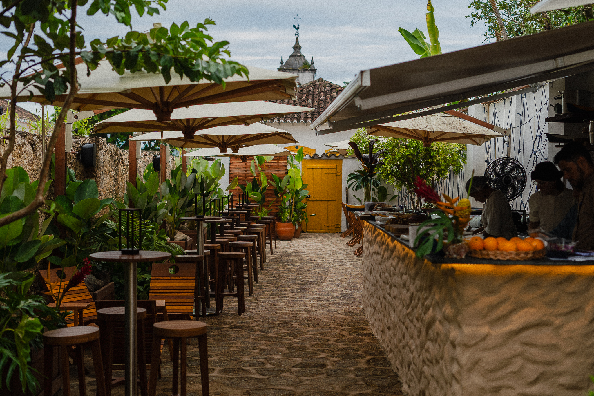 P&aacute;tio externo de restaurante com mesas e banquetas de madeira sob guarda-s&oacute;is brancos, plantas verdes e um balc&atilde;o de pedra com frutas e funcion&aacute;rios. Ao fundo, uma porta amarela e um telhado colonial com cata-vento