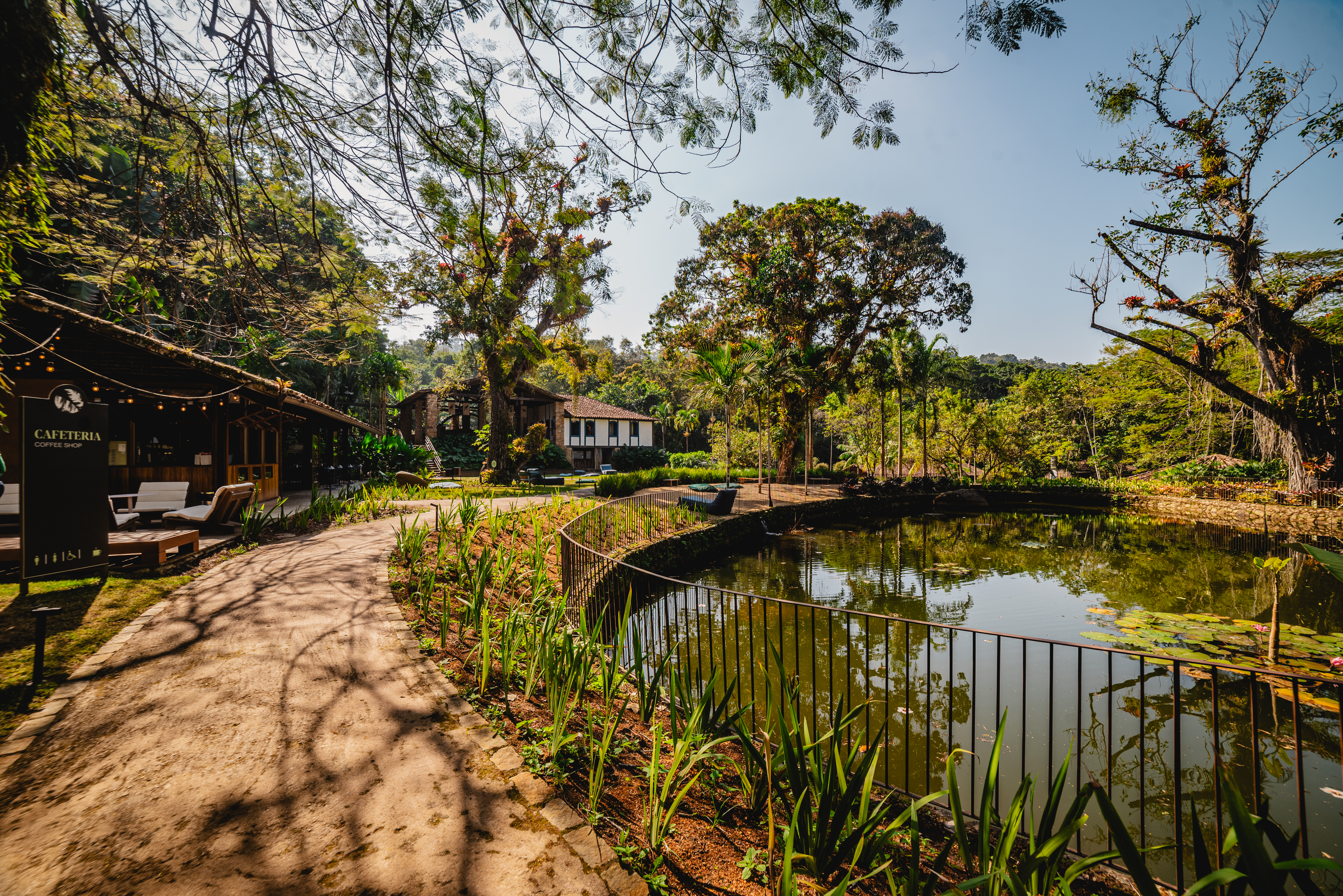 Caminho de terra batida ladeado por vegeta&ccedil;&atilde;o e um lago com plantas aqu&aacute;ticas, &agrave; direita. &Agrave; esquerda, uma cafeteria r&uacute;stica com letreiro CAFETERIA COFFEE SHOP e bancos de madeira. Ao fundo, casas e &aacute;rvores altas sob c&eacute;u azul