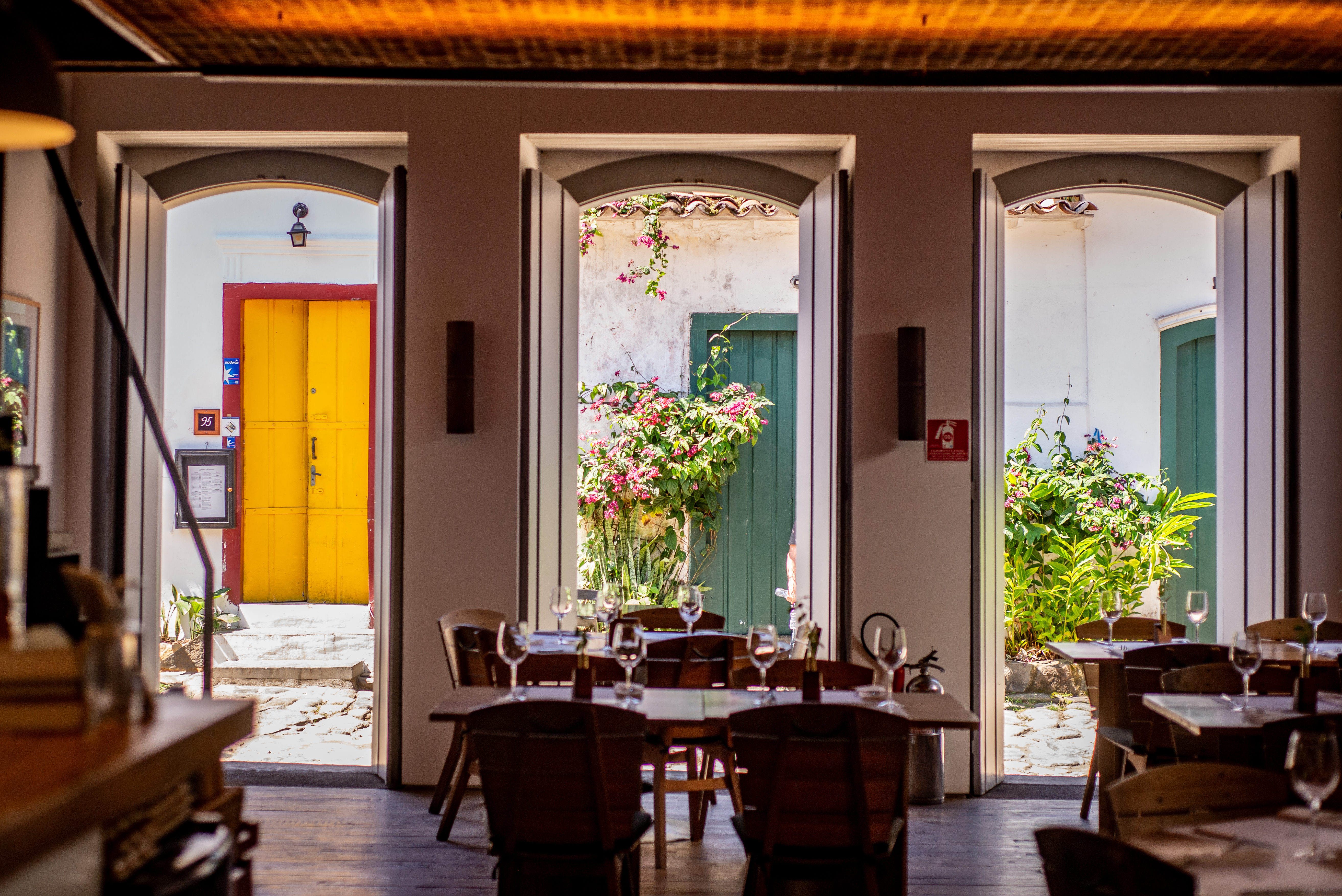 Interior de um restaurante com mesas de madeira e cadeiras escuras, com ta&ccedil;as de vinho. Tr&ecirc;s portas arqueadas revelam um p&aacute;tio externo com plantas e flores, e uma porta amarela vibrante ao fundo. O ch&atilde;o &eacute; de madeira escura e o teto tem um acabamento r&uacute;stico