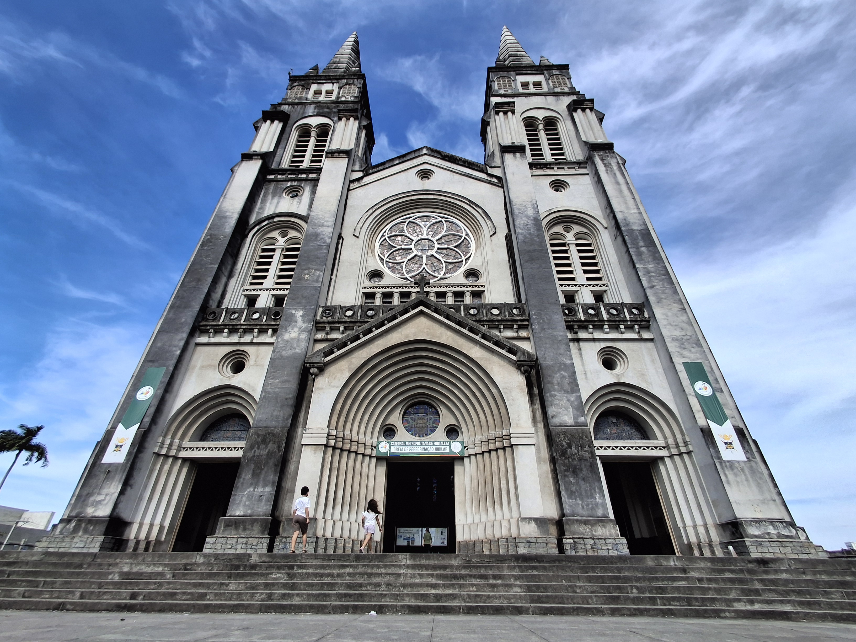 Fachada imponente da Bas&iacute;lica Santu&aacute;rio de Nazar&eacute;, em Bel&eacute;m, com duas torres pontiagudas e um grande ros&aacute;ceo central. Dois visitantes sobem os degraus em dire&ccedil;&atilde;o &agrave; entrada principal, sob um c&eacute;u azul com nuvens esparsas. Faixas verdes com s&iacute;mbolos religiosos adornam as laterais da constru&ccedil;&atilde;o cinza