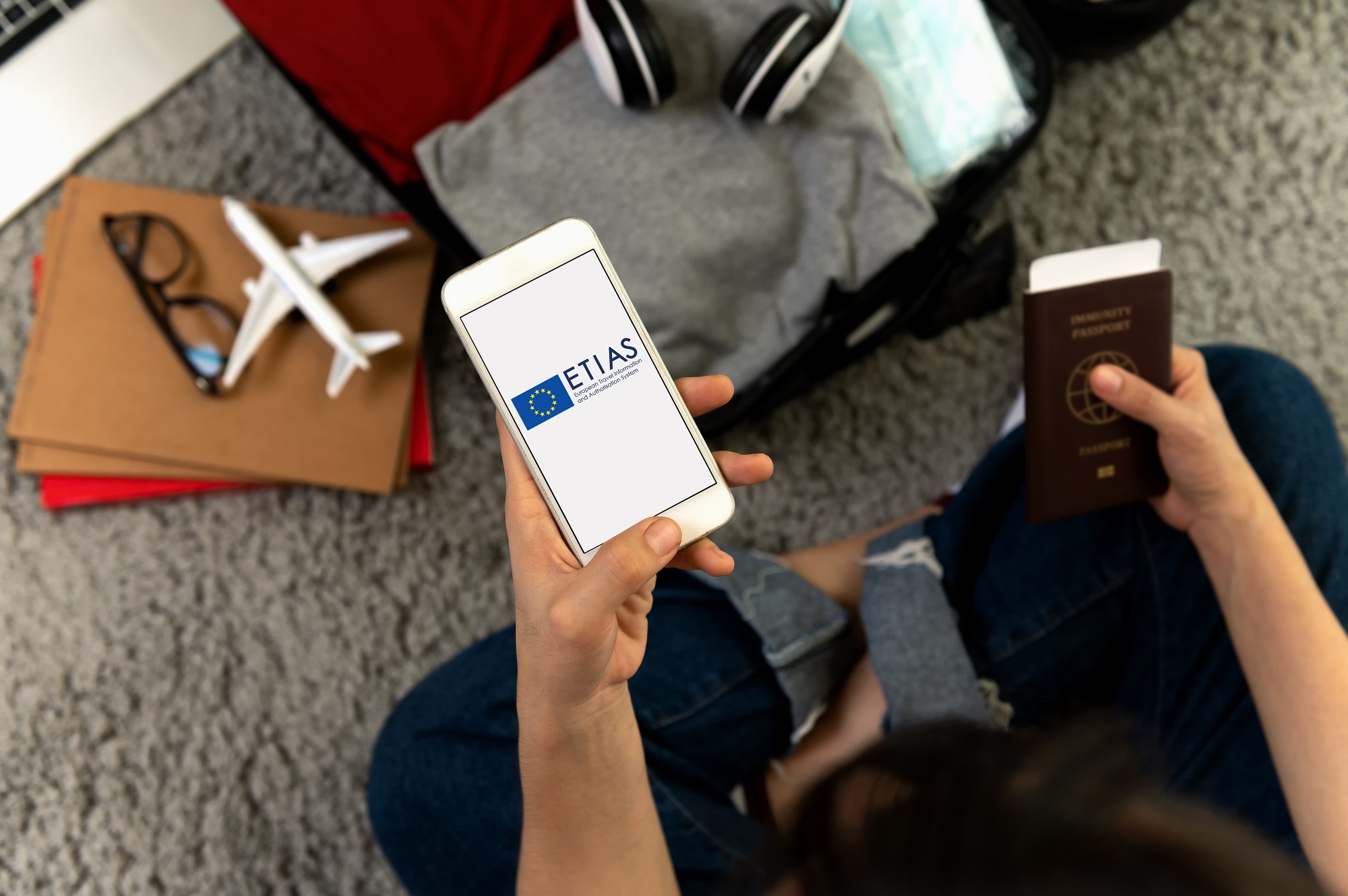 Pessoa sentada no chão com jeans, segurando um celular com o logo ETIAS e um passaporte. Ao lado, uma mala aberta com roupas, fones de ouvido, máscara, óculos e um avião em miniatura sobre cadernos, sugerindo preparativos para viagem