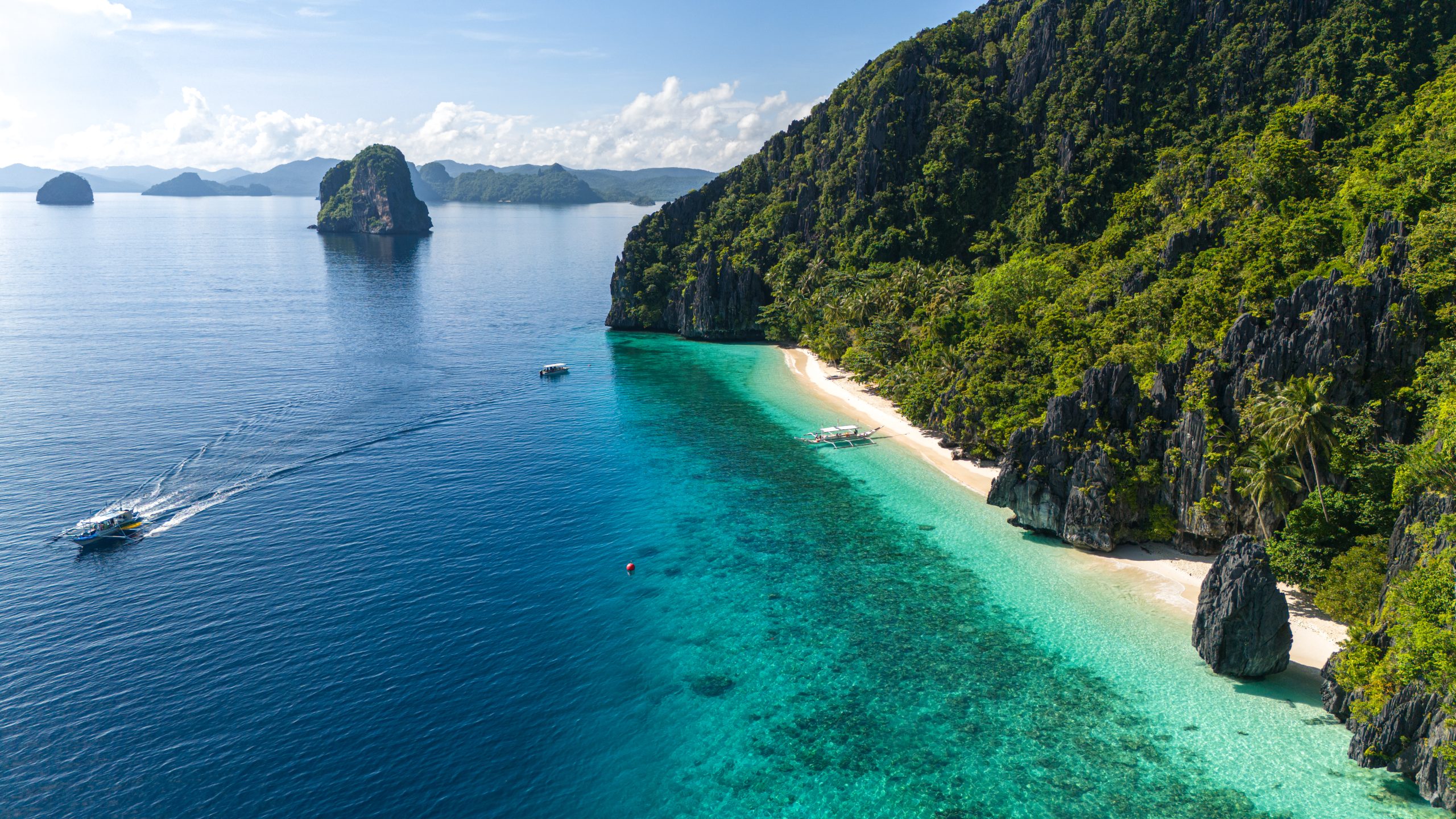 Vista aérea de uma praia paradisíaca com areia branca e águas cristalinas em tons de azul e verde. Uma lancha navega em mar aberto, enquanto barcos de passeio estão ancorados perto da costa. Montanhas rochosas cobertas por vegetação densa e palmeiras emolduram a paisagem sob um céu azul com nuvens esparsas