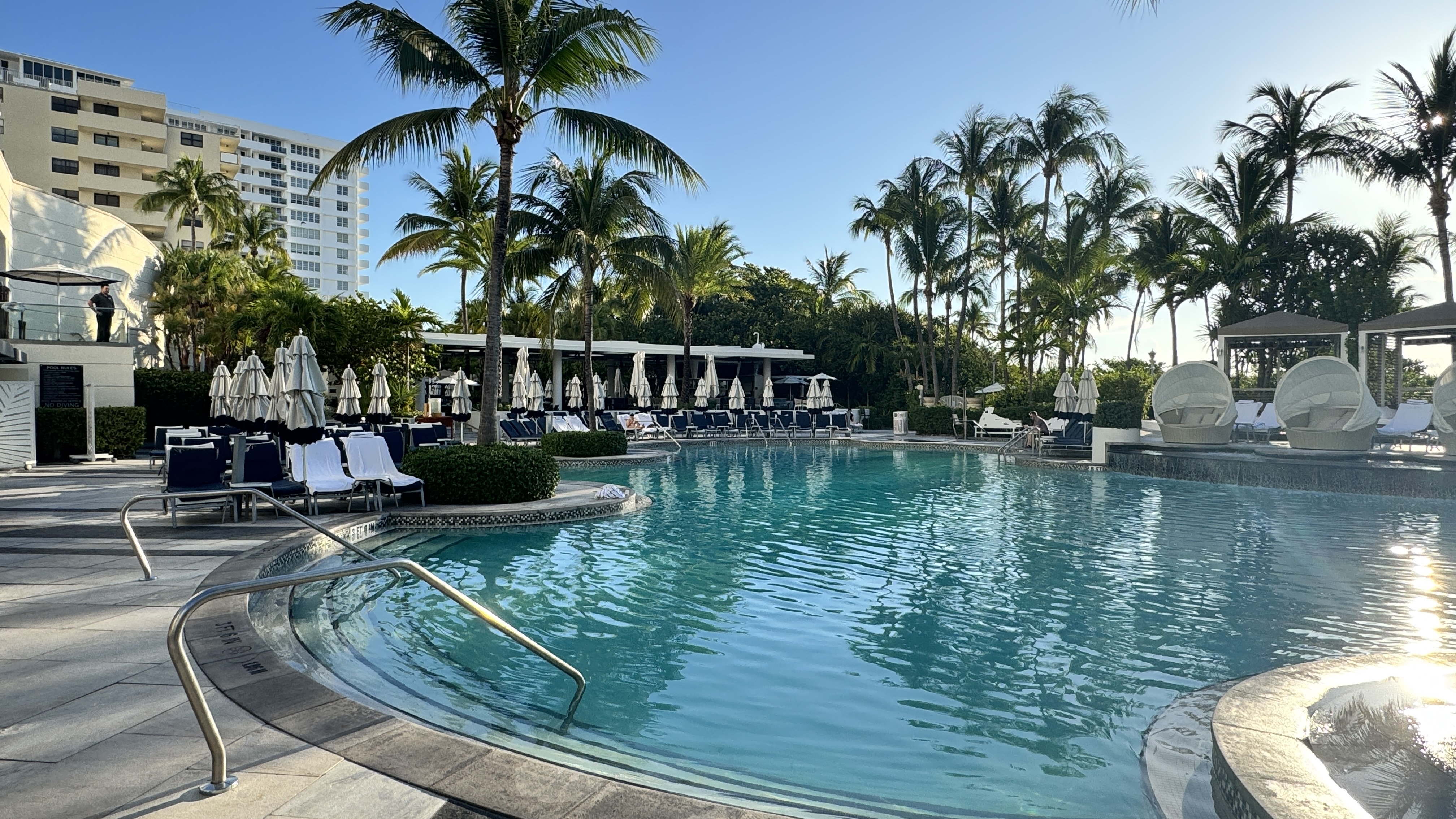Piscina de hotel com &aacute;gua azul-turquesa, rodeada por espregui&ccedil;adeiras, guarda-s&oacute;is brancos e palmeiras. Ao fundo, pr&eacute;dios altos e o sol refletindo na &aacute;gua, criando um ambiente de lazer e relaxamento. Um homem observa da varanda &agrave; esquerda