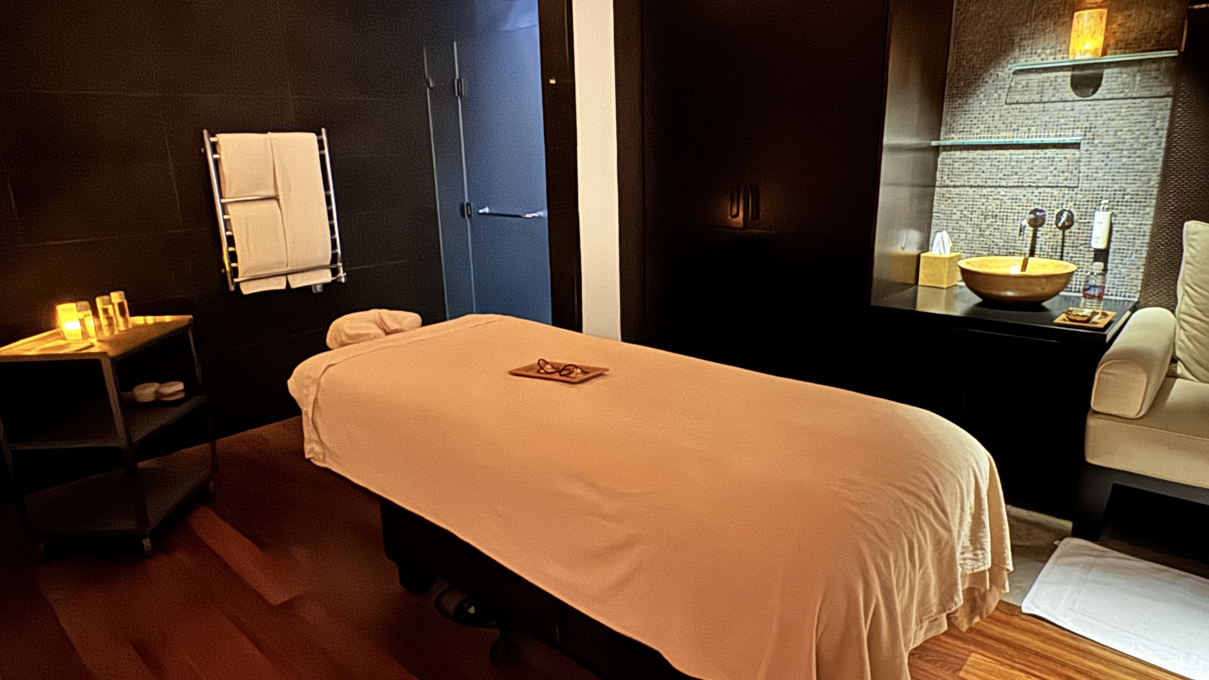 Sala de spa com ilumina&ccedil;&atilde;o suave, maca de massagem coberta por len&ccedil;ol p&ecirc;ssego, &oacute;culos sobre bandeja de madeira. &Agrave; esquerda, mesa de apoio com velas acesas e toalhas em aquecedor. &Agrave; direita, pia dourada sobre balc&atilde;o escuro e poltrona creme. O ambiente &eacute; relaxante e acolhedor