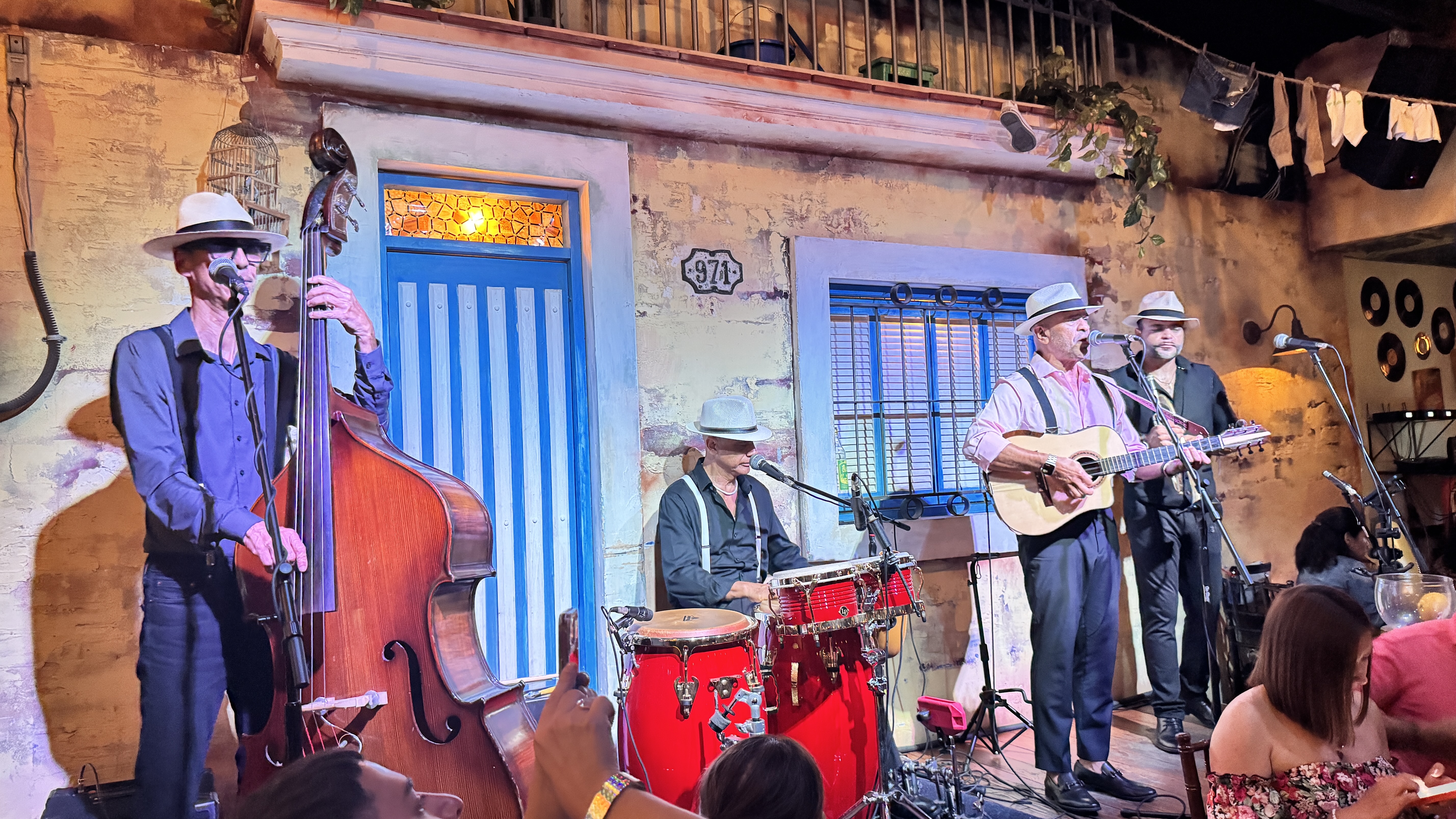Quatro m&uacute;sicos homens, todos de chap&eacute;u, tocam instrumentos em um palco decorado como uma fachada de casa antiga. Um toca contrabaixo, outro bateria, e dois viol&atilde;o, cantando em microfones. O p&uacute;blico assiste em primeiro plano