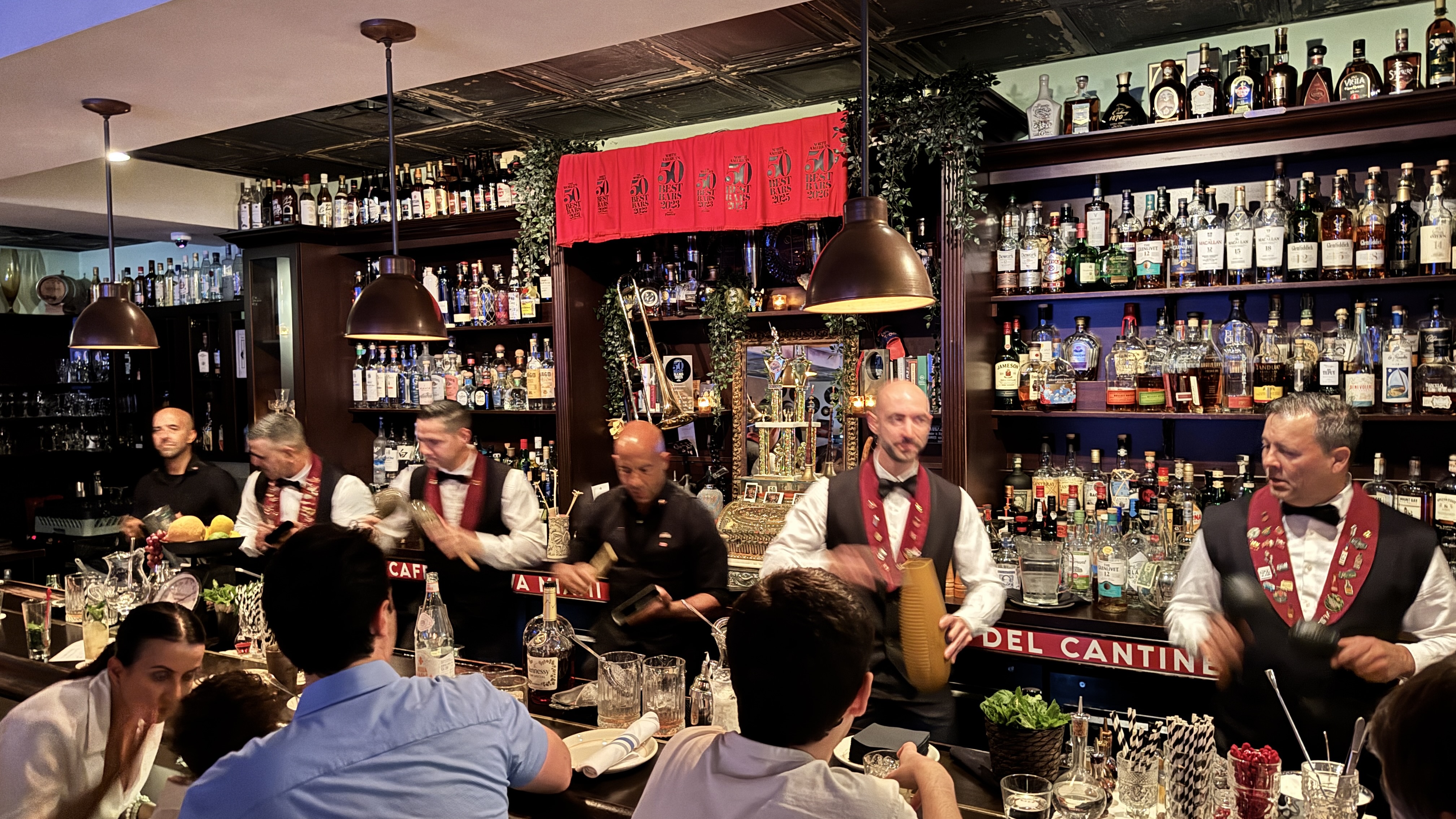 Seis bartenders, cinco homens e uma mulher, preparam bebidas em um bar com prateleiras cheias de garrafas de bebidas alco&oacute;licas. Dois homens usam coletes pretos com la&ccedil;os vermelhos e medalhas, enquanto os outros usam camisas pretas. Clientes sentam-se &agrave; frente do balc&atilde;o, observando a prepara&ccedil;&atilde;o. O ambiente &eacute; iluminado por pendentes de teto e decorado com folhagens e um espelho dourado. Uma faixa vermelha com caracteres dourados decora a parede.