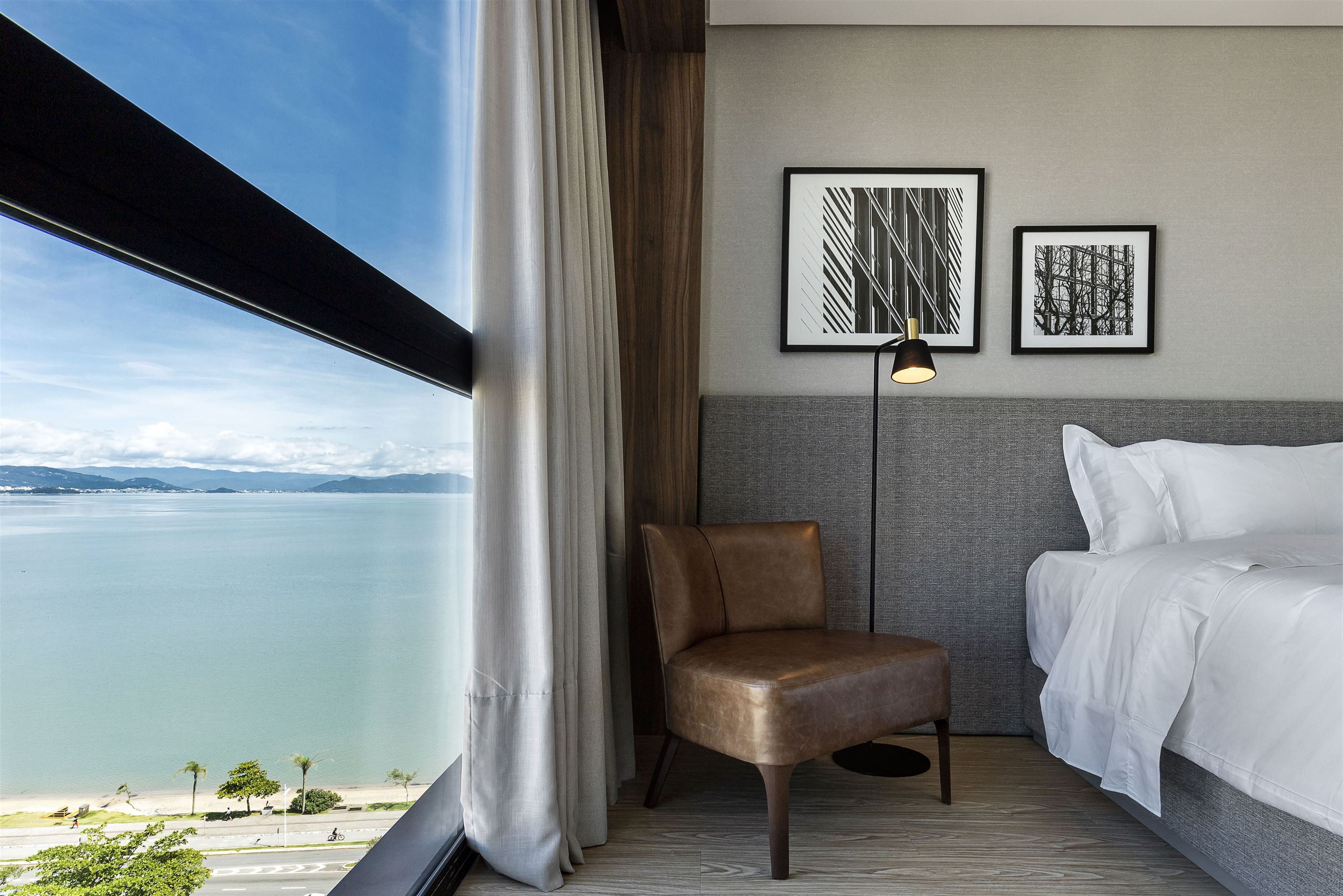Quarto de hotel moderno com vista panorâmica para o mar azul e montanhas distantes. À esquerda, uma janela ampla com cortina bege. À direita, uma cama com lençóis brancos, uma poltrona de couro marrom e um abajur de chão preto com detalhes dourados. Duas obras de arte abstratas em preto e branco decoram a parede cinza acima da cama