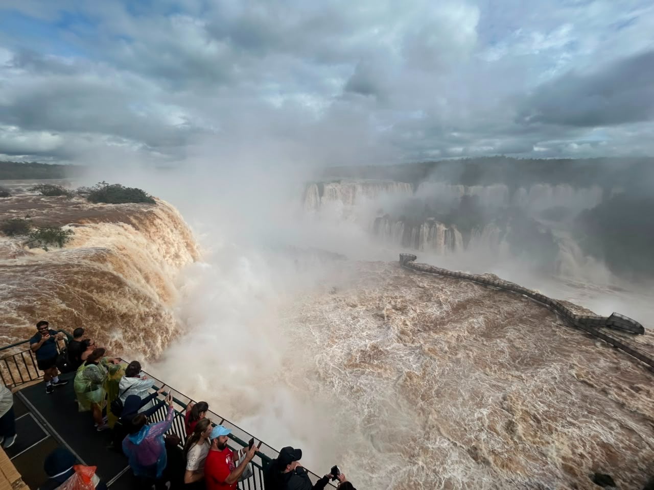 Vista aérea das Cataratas do Iguaçu com grande volume de água marrom e névoa intensa. Turistas observam a queda dágua de uma passarela à esquerda, muitos tirando fotos. O céu está nublado, e a vegetação densa da floresta aparece ao fundo. A passarela que leva à Garganta do Diabo está submersa