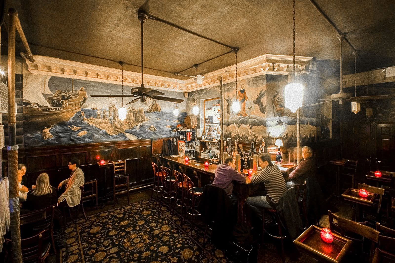 Interior de um bar com iluminação baixa e ambiente aconchegante. Murais com cenas marítimas e figuras clássicas decoram as paredes acima de painéis de madeira escura. No centro, um balcão com bancos vermelhos, onde três homens conversam. À esquerda, duas mulheres sentadas em uma mesa. O chão tem um tapete estampado e há velas acesas em mesas de madeira, criando uma atmosfera intimista