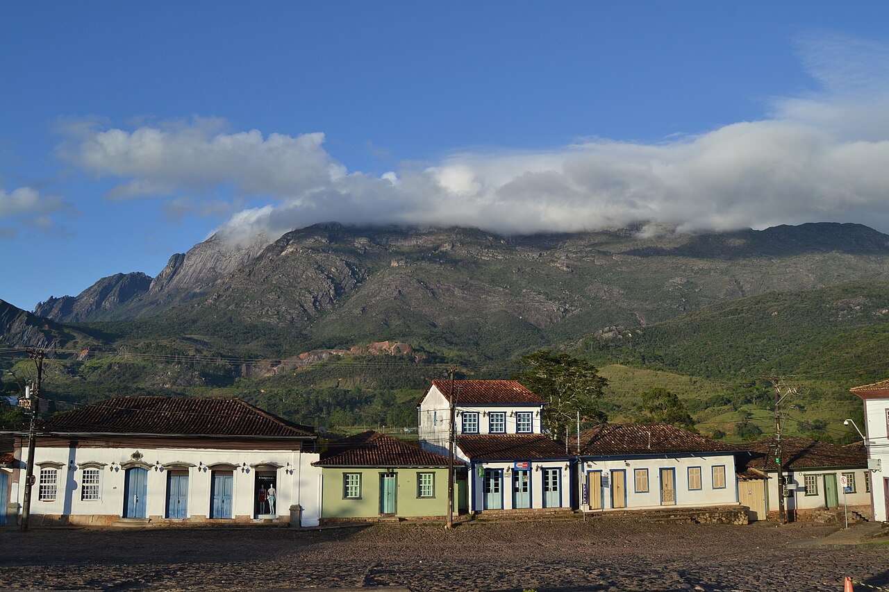 Casas coloniais coloridas em primeiro plano, com telhados de barro e janelas gradeadas, em uma rua de paralelepípedos. Ao fundo, montanhas rochosas e verdes, com nuvens baixas cobrindo os picos, sob um céu azul