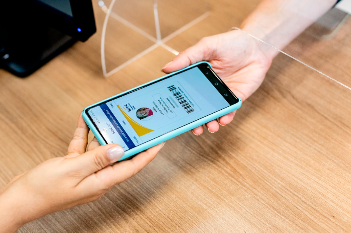 Duas mãos seguram um smartphone com capa azul-clara, exibindo um cartão digital do Sesc com foto e código de barras, sobre uma mesa de madeira clara