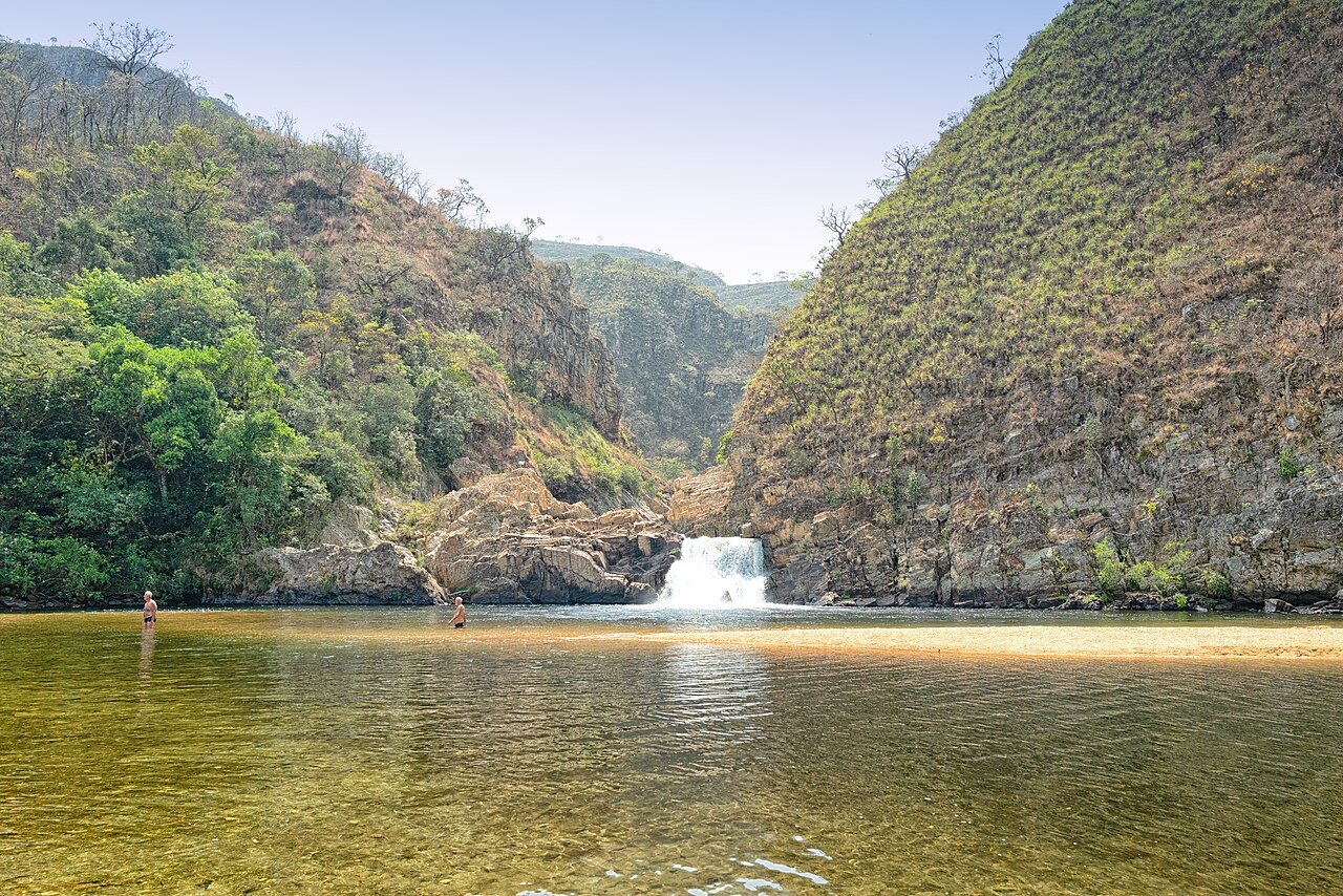 Duas pessoas em pé na água rasa de um rio de cor esverdeada, com uma cachoeira ao fundo entre montanhas rochosas e vegetação seca e verde. Foto sob licença CC BY 2.0
