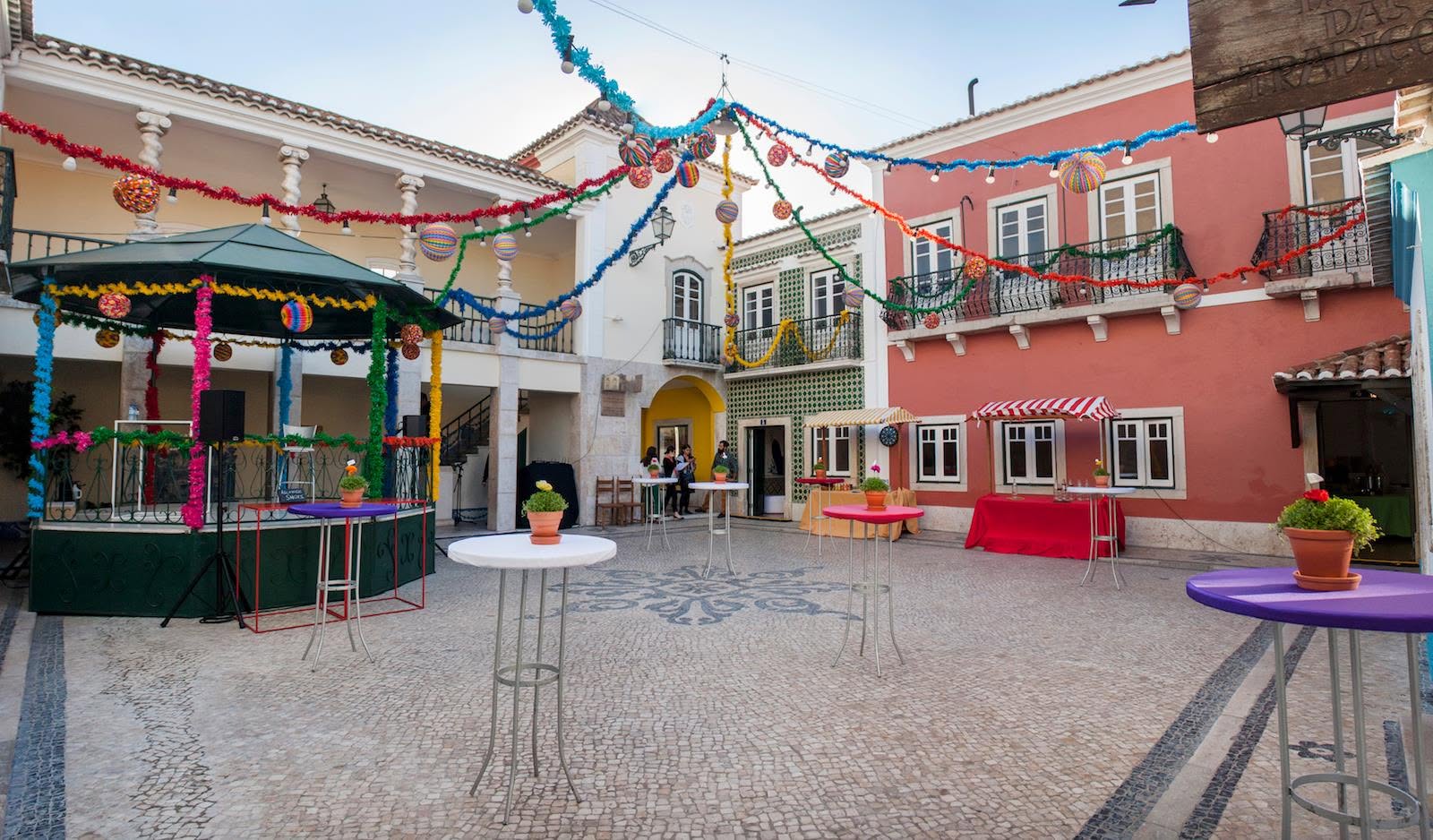 Pátio de paralelepípedos decorado para festa, com coreto verde, mesas altas coloridas e guirlandas vibrantes penduradas entre edifícios de fachadas brancas, vermelhas e azulejadas. Pessoas conversam ao fundo, e há um toldo listrado vermelho e branco.