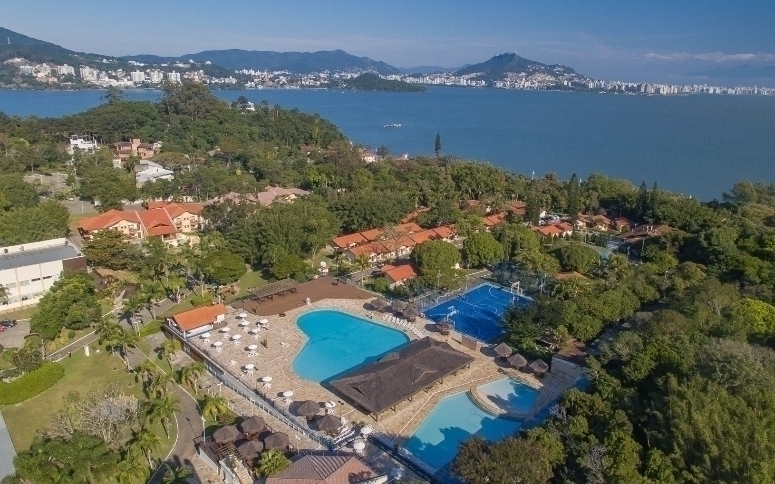 Vista aérea de um resort com piscinas azuis, quadra esportiva, bangalôs e telhados de cerâmica, cercado por vegetação exuberante. Ao fundo, um mar azul e uma cidade costeira com montanhas sob céu claro