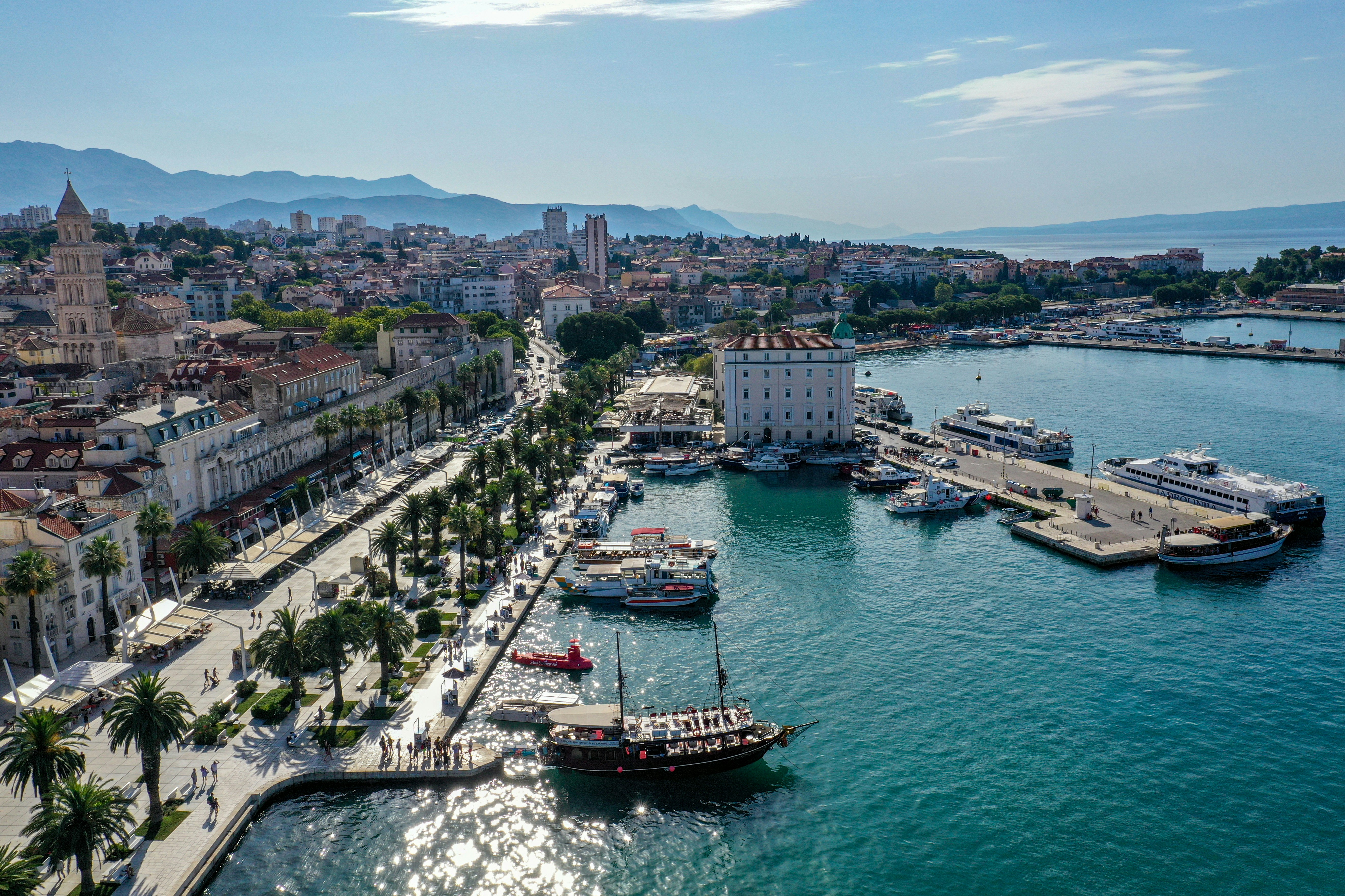 Vista aérea de uma cidade costeira com prédios históricos, um campanário alto e um calçadão com palmeiras. Barcos e navios de diferentes tamanhos estão atracados no porto, com montanhas ao fundo sob um céu azul claro