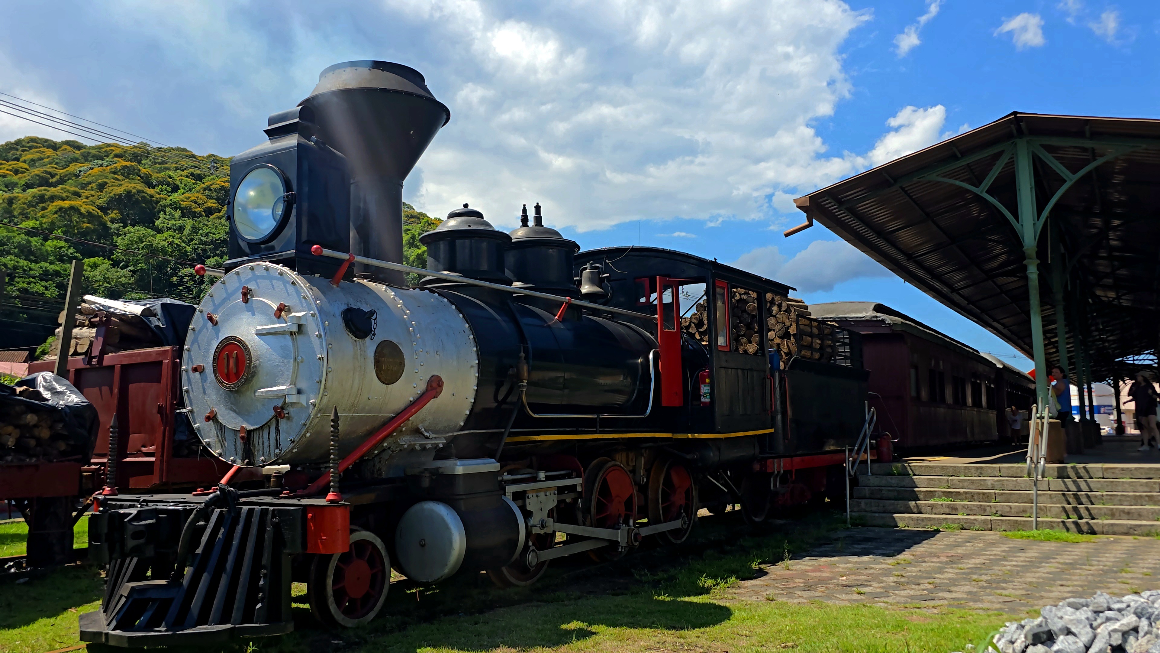Locomotiva a vapor preta e prateada, com detalhes vermelhos, estacionada em um gramado ao lado de uma esta&ccedil;&atilde;o de trem antiga com telhado verde e marrom, sob c&eacute;u azul com nuvens brancas