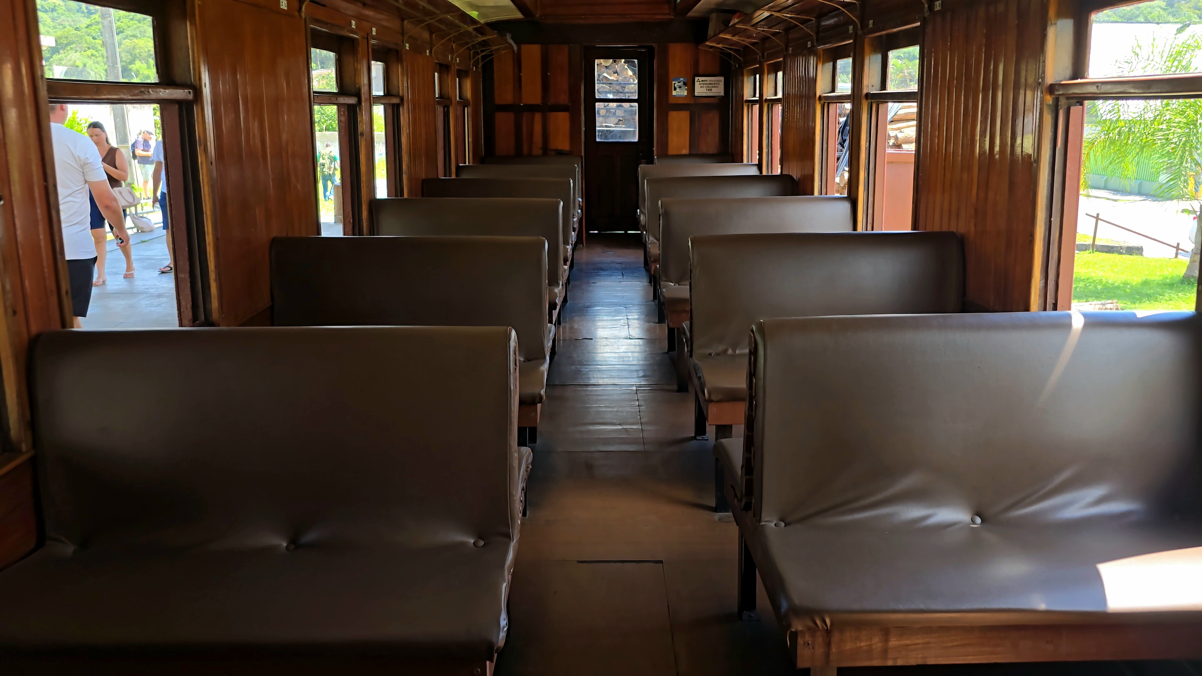 Interior de um vag&atilde;o de trem antigo com paredes e teto de madeira, bancos estofados marrons dispostos em duas fileiras. Pessoas em p&eacute; s&atilde;o vis&iacute;veis atrav&eacute;s das janelas e portas abertas, com vegeta&ccedil;&atilde;o verde ao fundo.