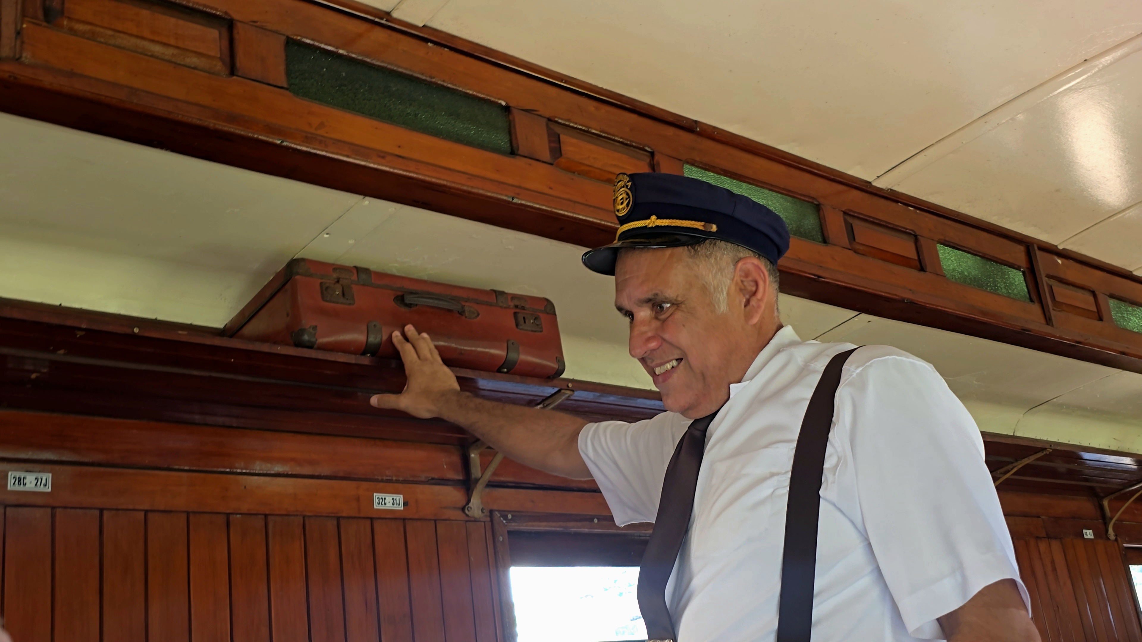 Homem de meia-idade, com uniforme de maquinista, sorri enquanto toca uma mala de couro marrom no bagageiro de um trem antigo com acabamento em madeira
