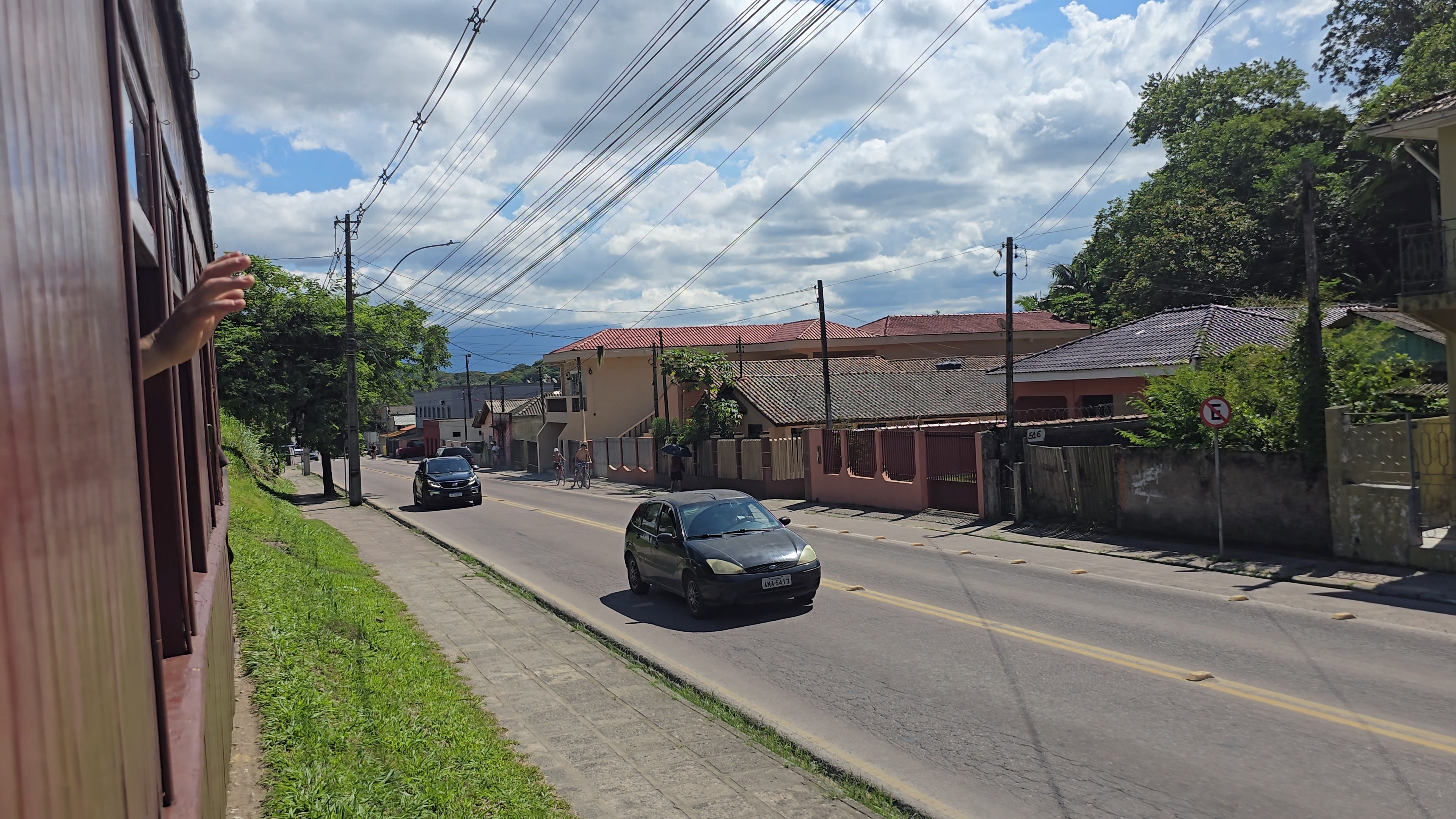 M&atilde;o de uma pessoa acenando pela janela de um trem em movimento, com uma rua paralela &agrave; direita, carros, casas e vegeta&ccedil;&atilde;o sob c&eacute;u azul com nuvens