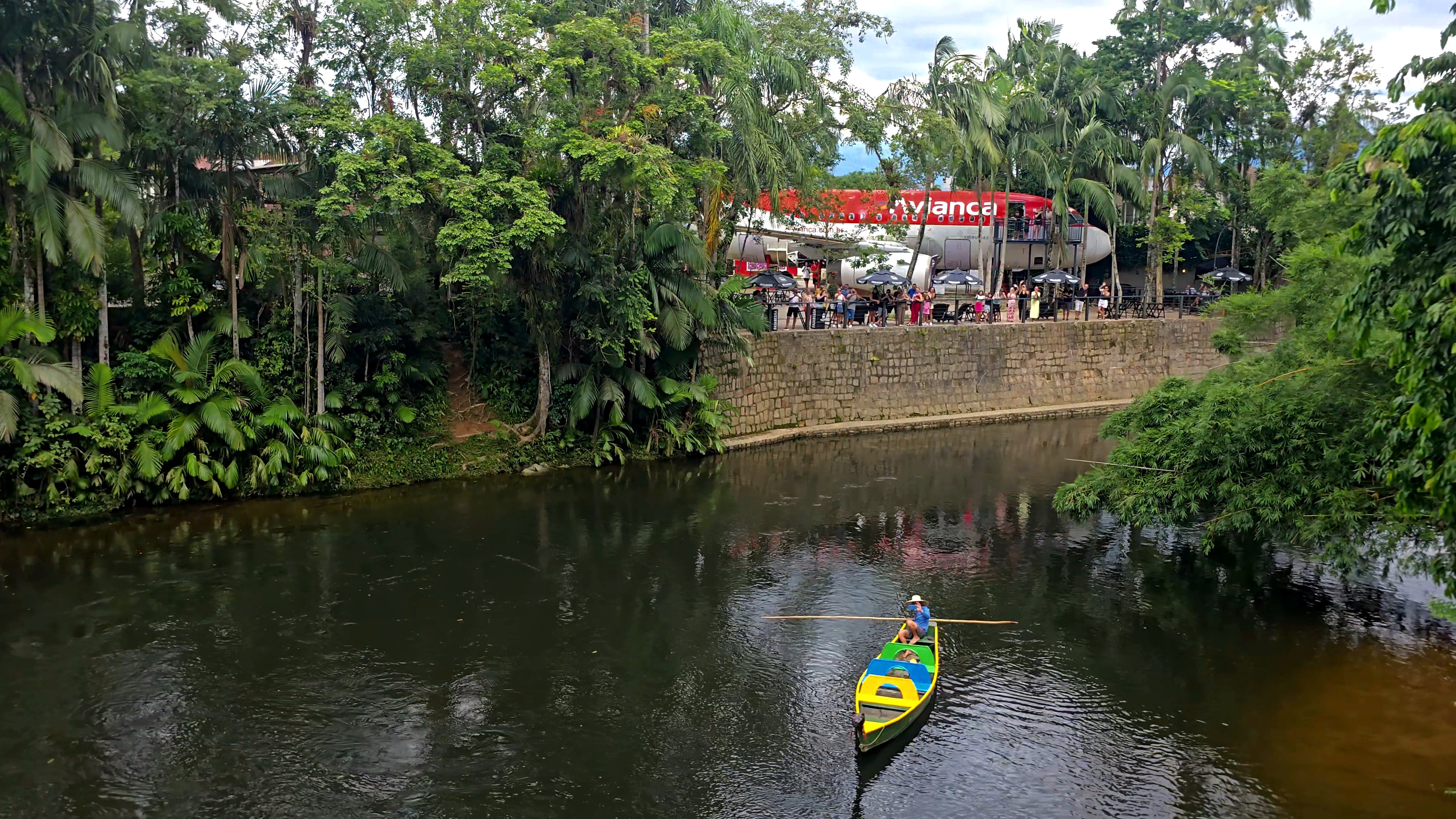 Um homem em um chap&eacute;u de palha rema um barco colorido (amarelo, azul e verde) em um rio escuro. &Agrave; direita, um muro de pedra sustenta uma &aacute;rea com pessoas e a fuselagem de um avi&atilde;o vermelho e branco da Avianca, cercado por densa vegeta&ccedil;&atilde;o tropical