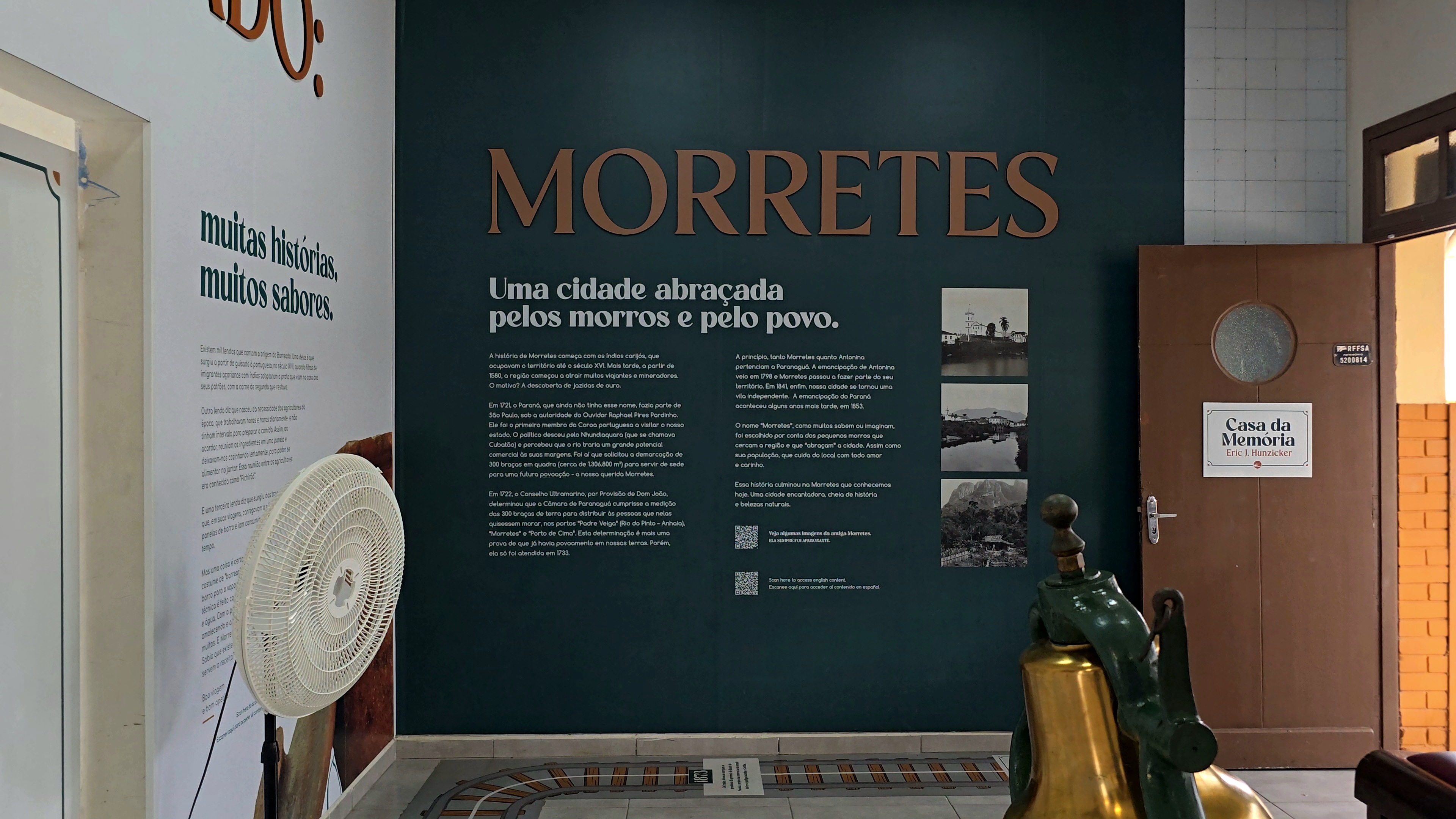 Parede verde-escura com texto MORRETES Uma cidade abra&ccedil;ada pelos morros e pelo povo e tr&ecirc;s fotos em preto e branco de paisagens. &Agrave; esquerda, parede branca com texto muitas hist&oacute;rias, muitos sabores e um ventilador branco. &Agrave; direita, porta marrom com placa Casa da Mem&oacute;ria. No ch&atilde;o, trilhos de trem em miniatura e um sino dourado e verde