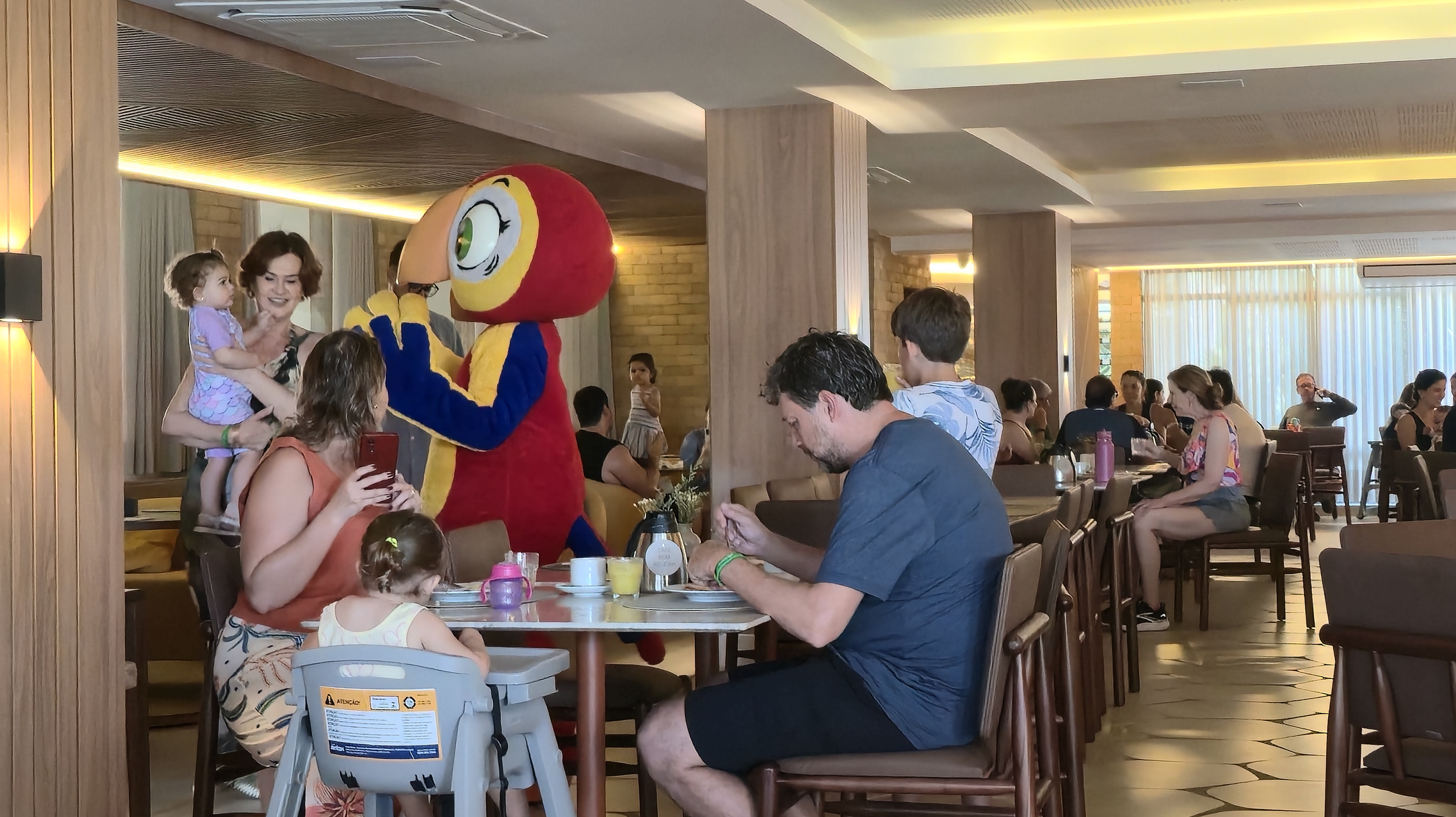 Pessoas em um restaurante com um mascote de papagaio vermelho e amarelo. Uma mulher segura um beb&ecirc;, outra tira foto com celular, e um homem come &agrave; mesa.