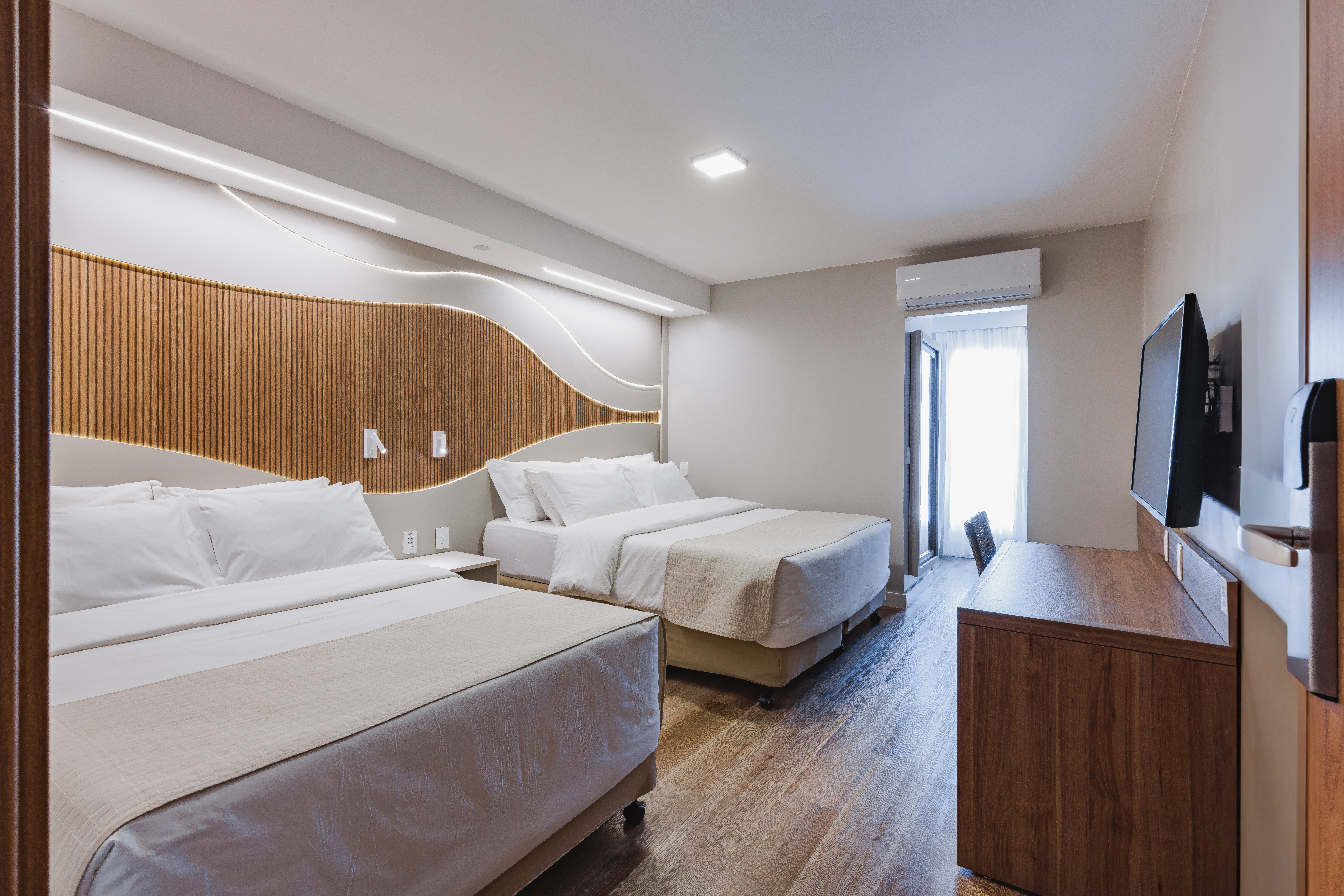 Quarto de hotel moderno com duas camas de casal, len&ccedil;&oacute;is brancos e colchas bege. A cabeceira tem pain&eacute;is de madeira verticais e ilumina&ccedil;&atilde;o embutida. &Agrave; direita, uma TV preta montada na parede acima de uma escrivaninha de madeira escura. Uma porta-janela aberta revela uma cortina branca e luz natural. O piso &eacute; de madeira clara