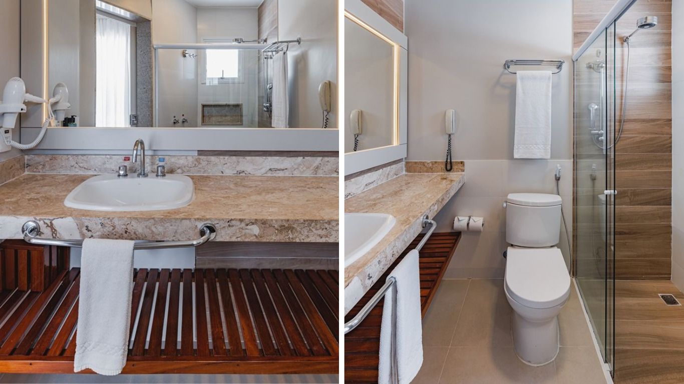 Banheiro moderno com bancada de m&aacute;rmore bege, pia branca, espelho grande e secador de cabelo. &Agrave; direita, vaso sanit&aacute;rio branco e box de vidro com chuveiro, parede revestida em madeira. Toalhas brancas penduradas em barras met&aacute;licas.