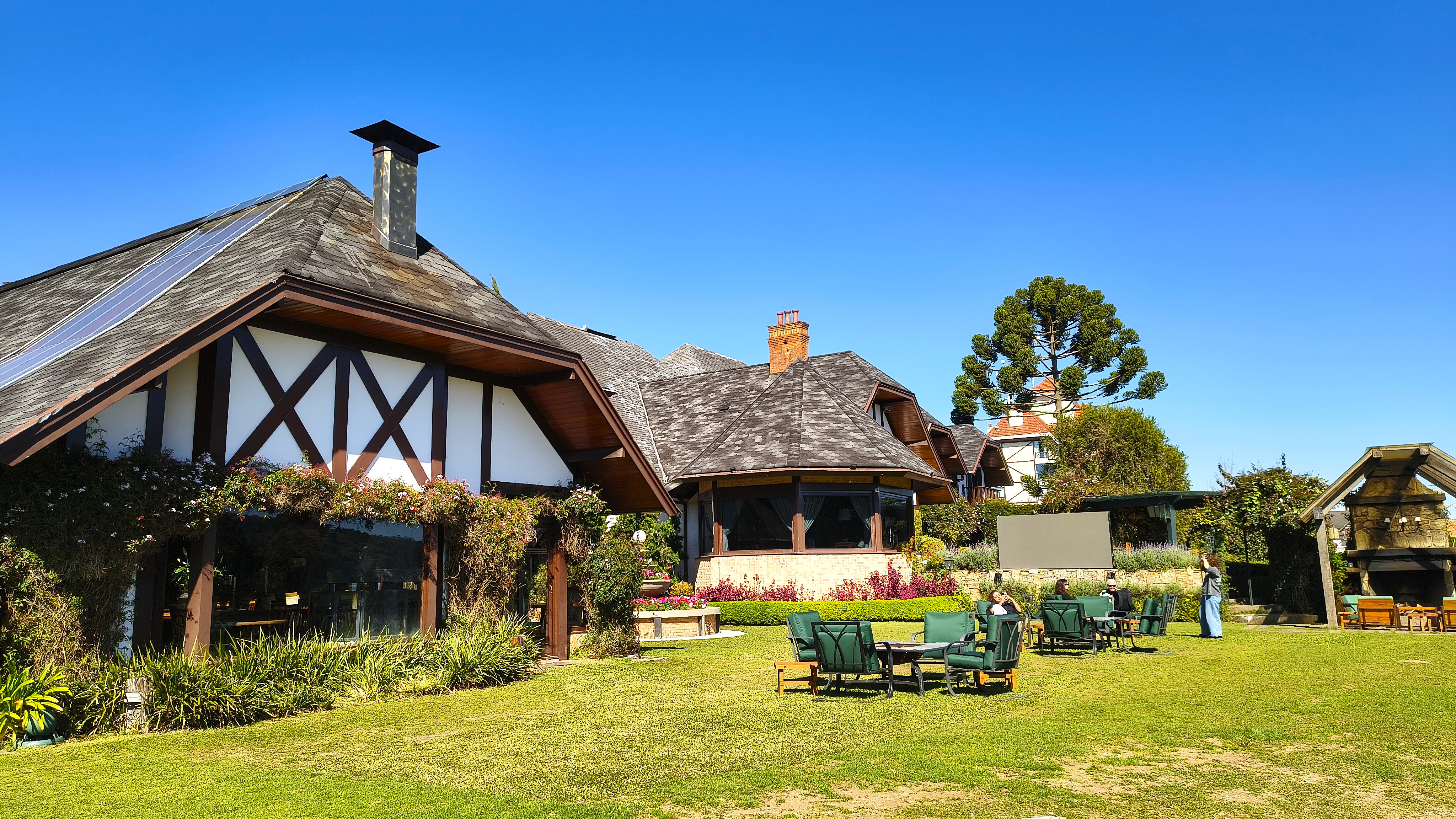 Casas com telhados de madeira e painéis solares, em um gramado verde com mesas e cadeiras de jardim, sob céu azul claro. Uma araucária alta se destaca ao fundo, e pessoas sentadas desfrutam do ambiente externo.