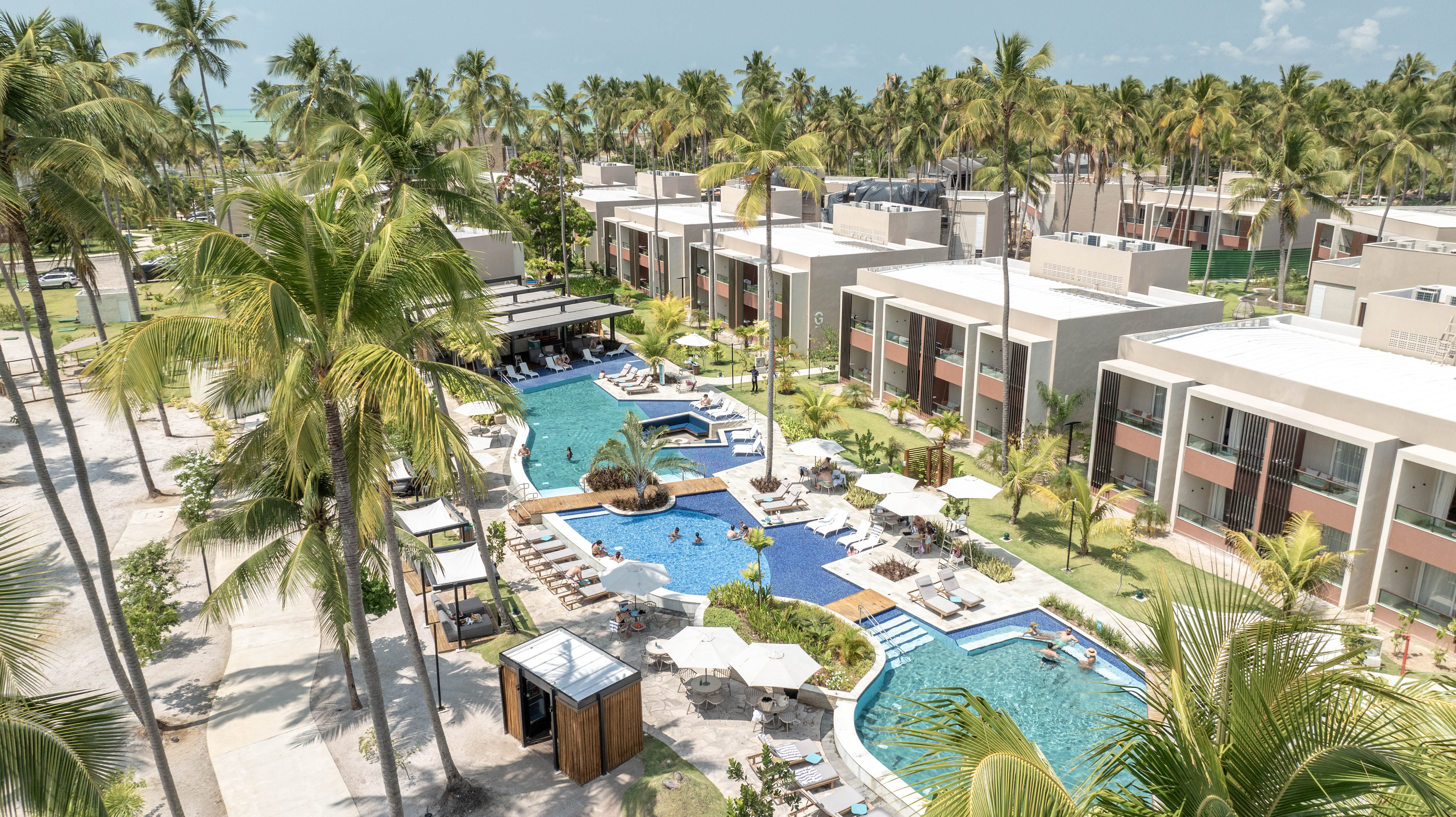 Vista aérea de um resort tropical com piscinas azuis sinuosas, cercadas por coqueiros altos e prédios de apartamentos modernos de dois andares com varandas. Pessoas nadam e relaxam em espreguiçadeiras sob guarda-sóis brancos, em um dia ensolarado